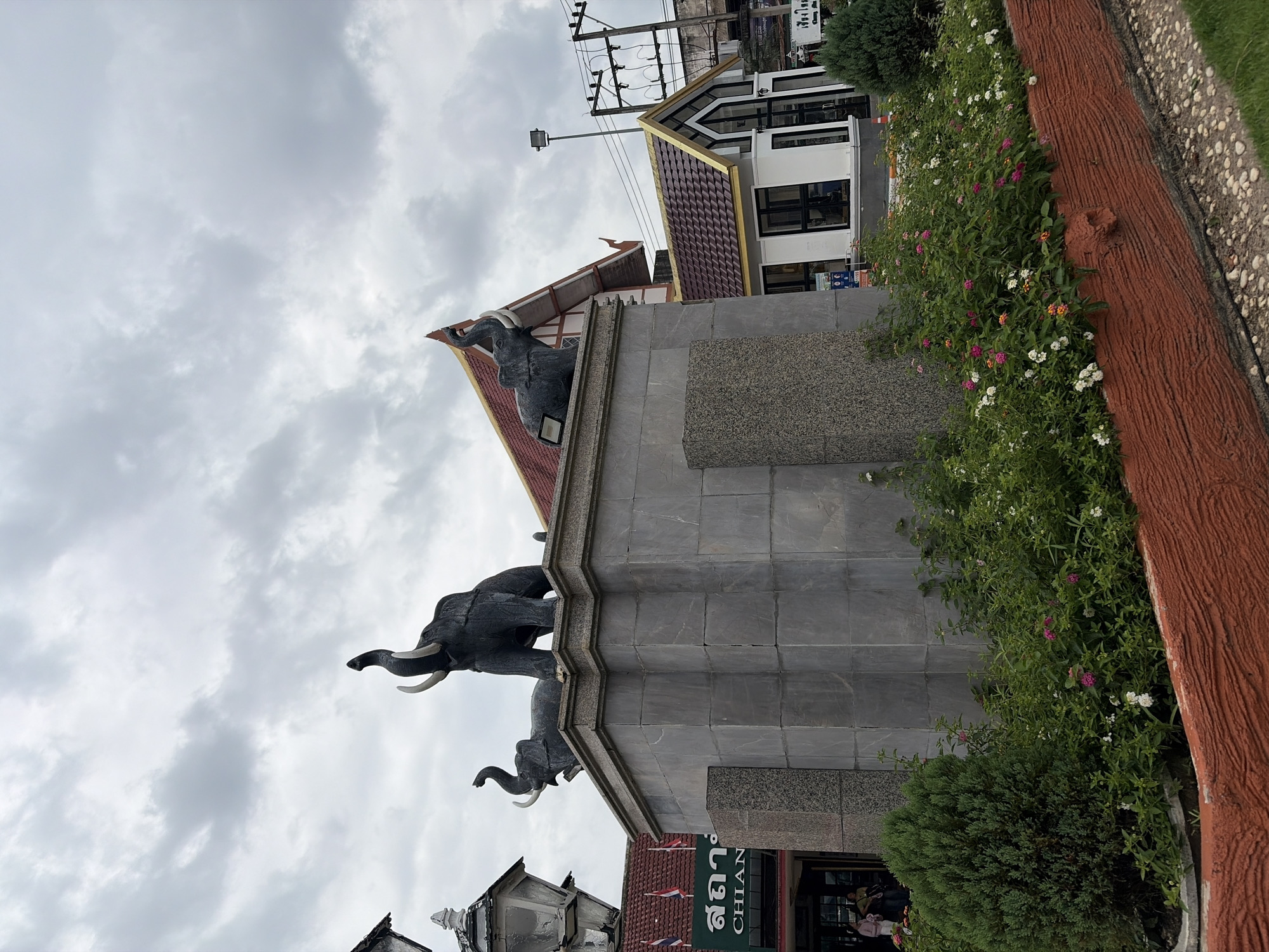 In Chiang Mai City Municipality, Thailand, a stone monument adorned with three dark elephant statues stands prominently. The central and largest elephant is positioned on the right, its trunk raised high, while a smaller elephant is to its left, also with an upward-raised trunk. Another smaller elephant is situated further left, with its trunk curled downwards. The monument's base is a grey, stone-clad structure, surrounded by lush green bushes and flowering plants in shades of pink, white, and orange. Behind the monument, a building with a distinctive red, multi-tiered roof and gold trim is visible. A smaller, modern white building with a similar red and gold roof is located to the right of the main building, featuring large glass windows. Electrical wires and poles are present in the background, indicating an urban setting. The sky is overcast with grey clouds. A few people are visible in the background, near the entrance of a building with Thai signage, including the word "CHIANG" on a green sign. A lamppost is situated to the left of the monument.