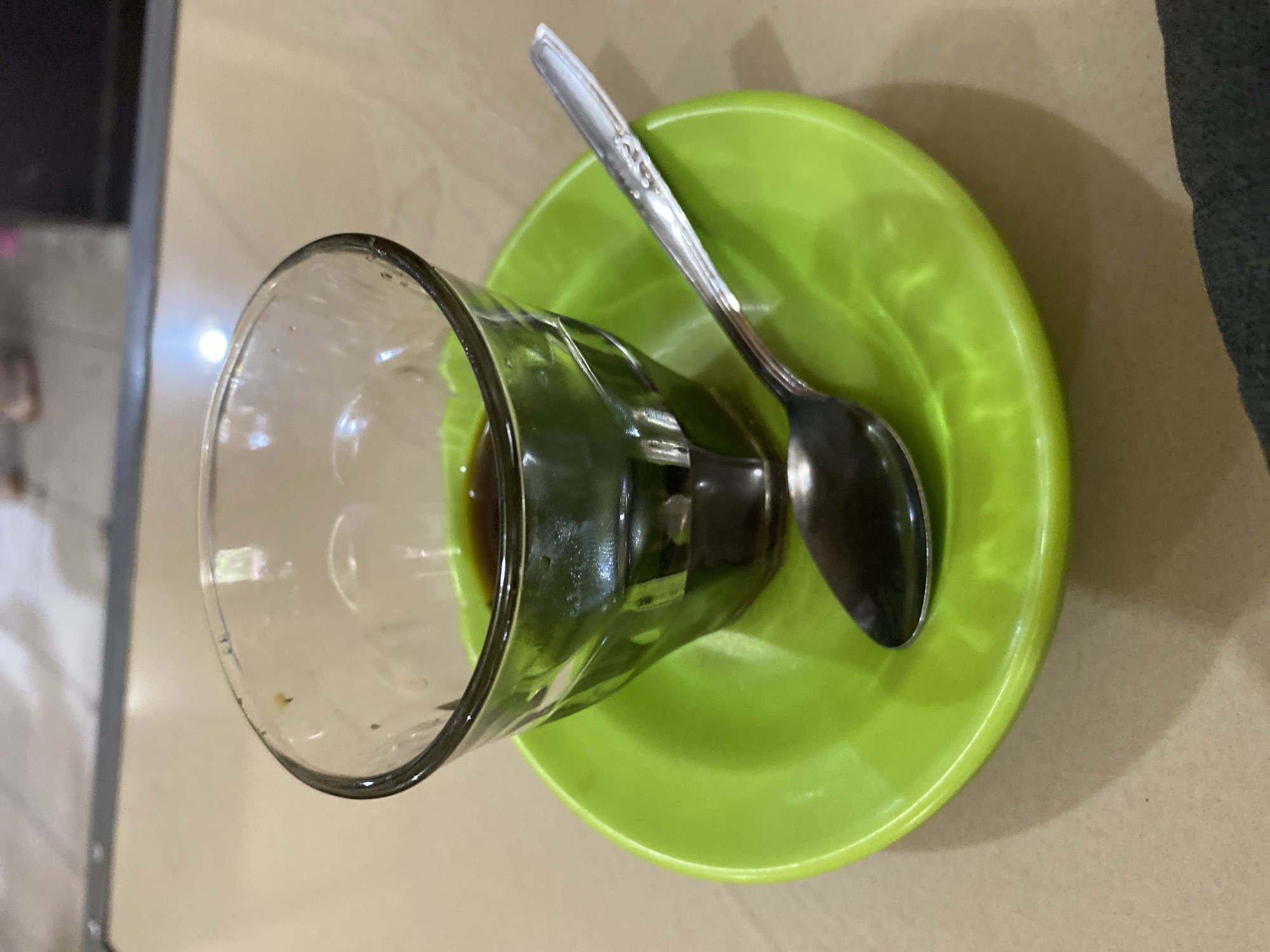A close-up, eye-level shot shows a small glass of dark coffee sitting on a bright green saucer. A silver spoon rests on the saucer, its bowl slightly submerged in the coffee. The glass has a clear, ribbed texture, and the coffee fills about a third of it. The saucer is smooth and circular, with a slight lip. The background is a blurred beige surface, likely a table, with a hint of flooring and possibly the legs of a chair or people in the far distance. This scene is likely from a cafe or restaurant in Lhokseumawe, Indonesia, given the common practice of serving coffee in such small glasses in that region. The lighting is subdued, with some reflections on the glass and saucer. No individuals are clearly discernible in the immediate vicinity of the coffee.