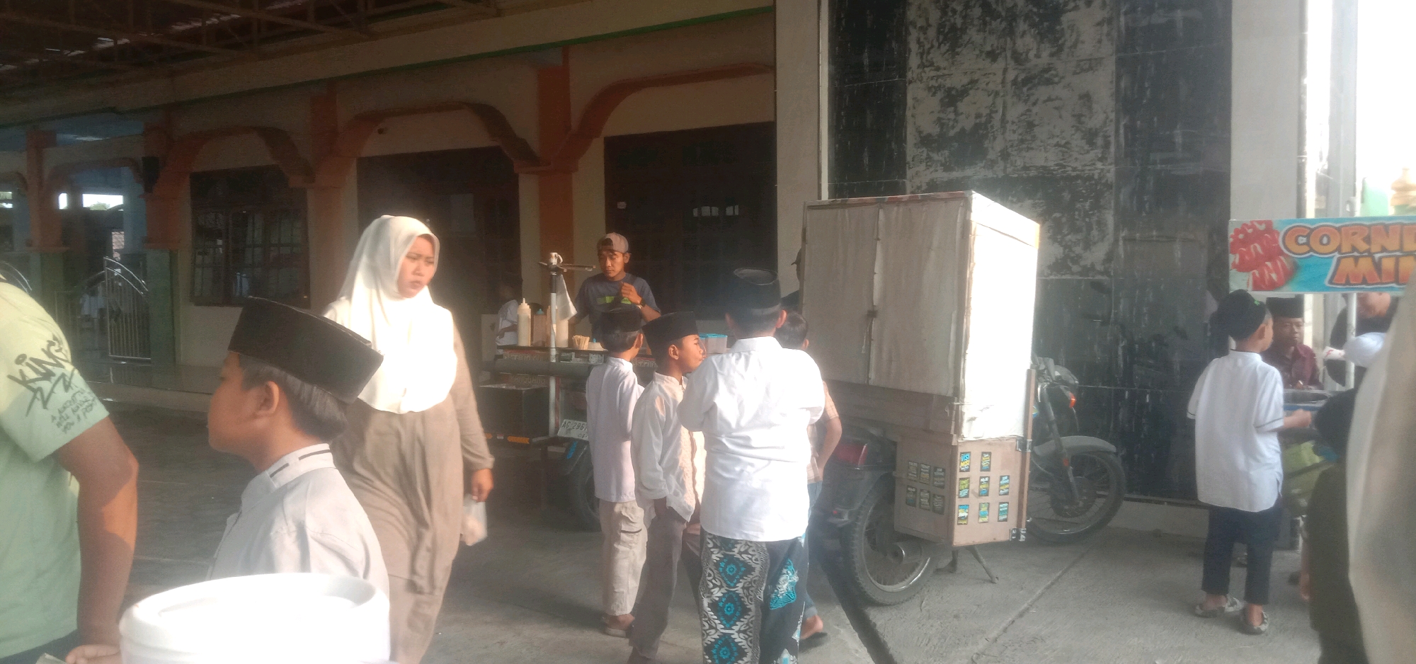 The image shows a street scene in Karang Dagangan, Indonesia. A group of people, including children wearing peci caps and adults, are gathered around a food cart. The cart appears to be selling drinks, with several bottles and dispensers visible. One person, possibly the vendor, is standing behind the cart. A woman in a white hijab is walking away from the cart, carrying a bag. In the background, there is a building with arched windows and a tiled roof. A sign with the words "CORNER MI" is visible on the right side of the image. The ground is paved, and there is a motorcycle parked next to the food cart. The lighting suggests it is daytime, possibly late afternoon, with sunlight casting shadows.
