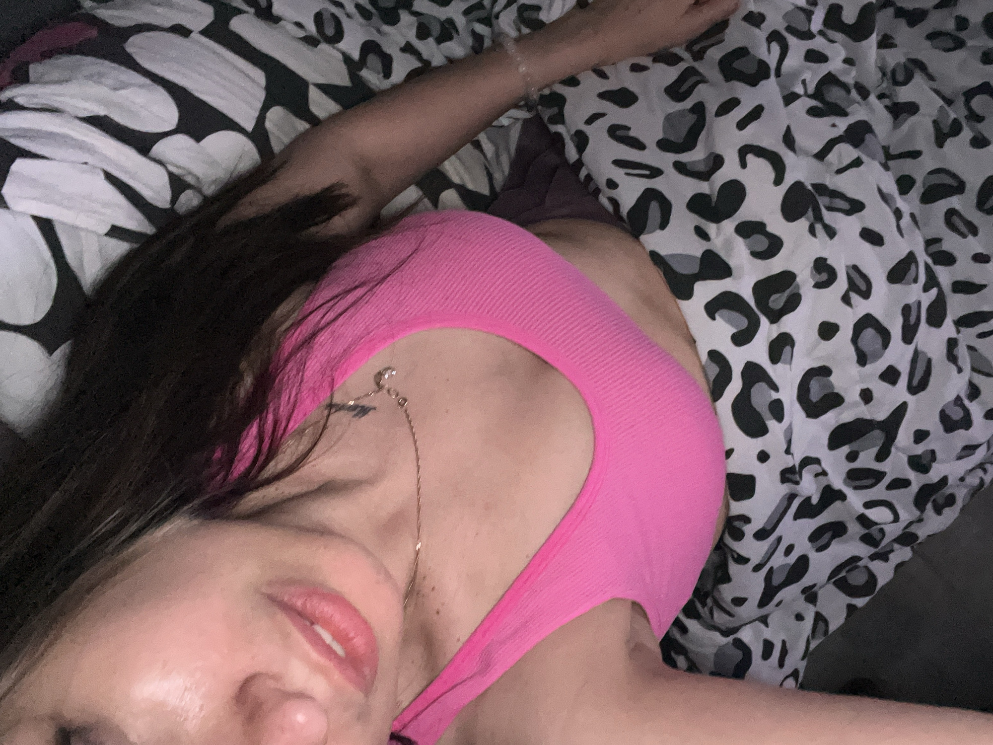 A woman is lying on a bed in an unknown location. She is wearing a pink top. The bed has two different patterned sheets, one with black and white shapes and the other with a black and white animal print.