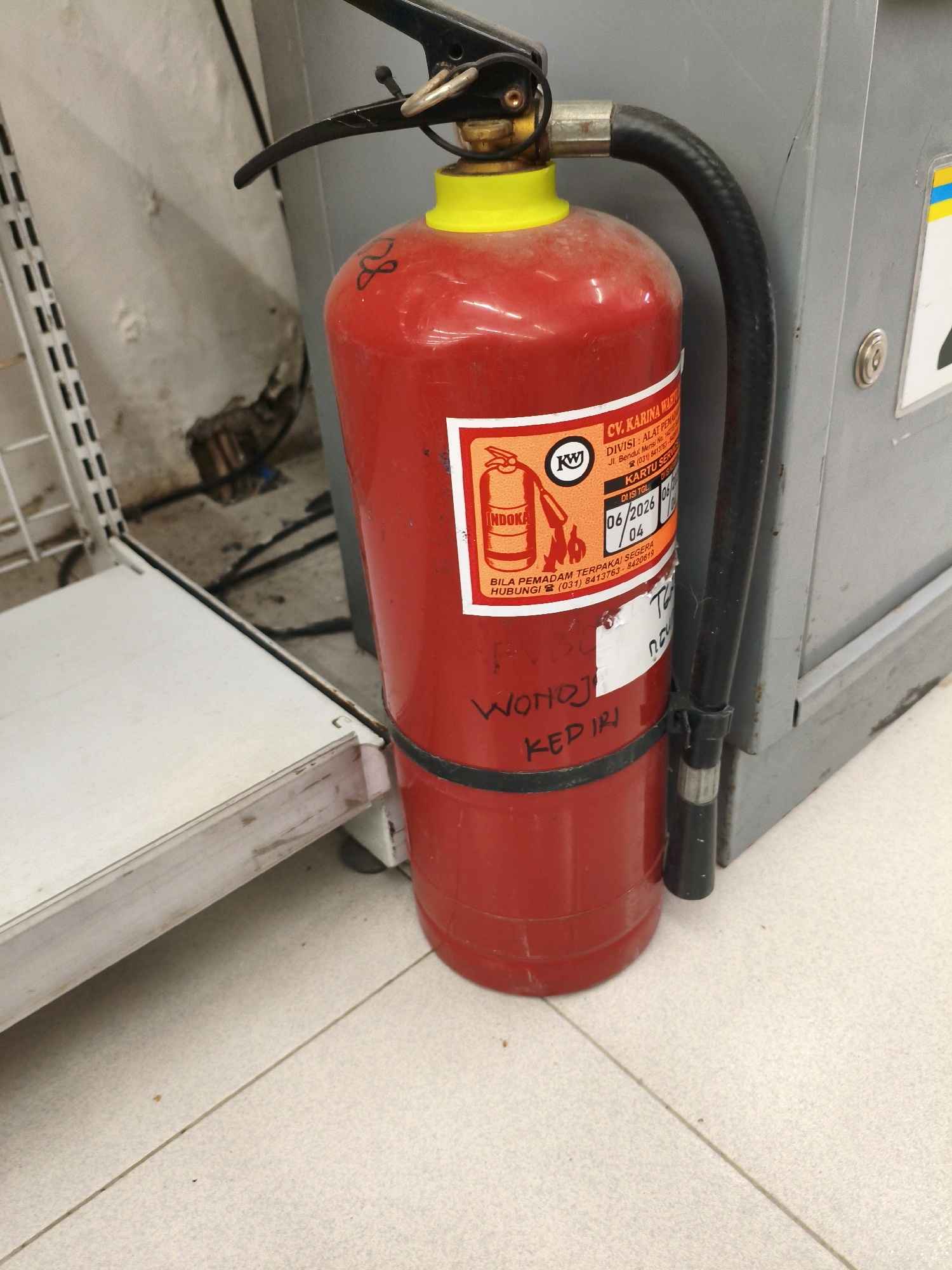 A red fire extinguisher with a yellow cap stands on a tiled floor in what appears to be a commercial setting. The fire extinguisher has a black hose attached to it and a label with Indonesian text and graphics, including a date stamp of "06/2026". Below the label, handwritten text reads "WONOKIRI" and "KEPIRI". To the left of the extinguisher is a white metal shelf unit, and to the right is a gray metal cabinet or appliance. The background wall is a light color and appears slightly worn or textured. The overall lighting is moderate. The scene is devoid of people or any visible action.