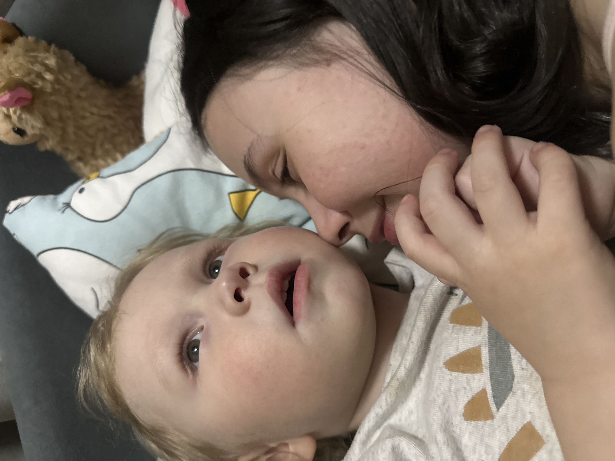A woman with dark hair and fair skin is lying down next to a young child with blond hair. The child is looking up at the camera with an open mouth. The woman is turned towards the child, holding its hand. A stuffed animal is visible in the upper left corner of the image. The background includes a pillow with a blue and white pattern. The location is not discernible from the image, and there is no information to suggest it is in Votkinsk, Russia.