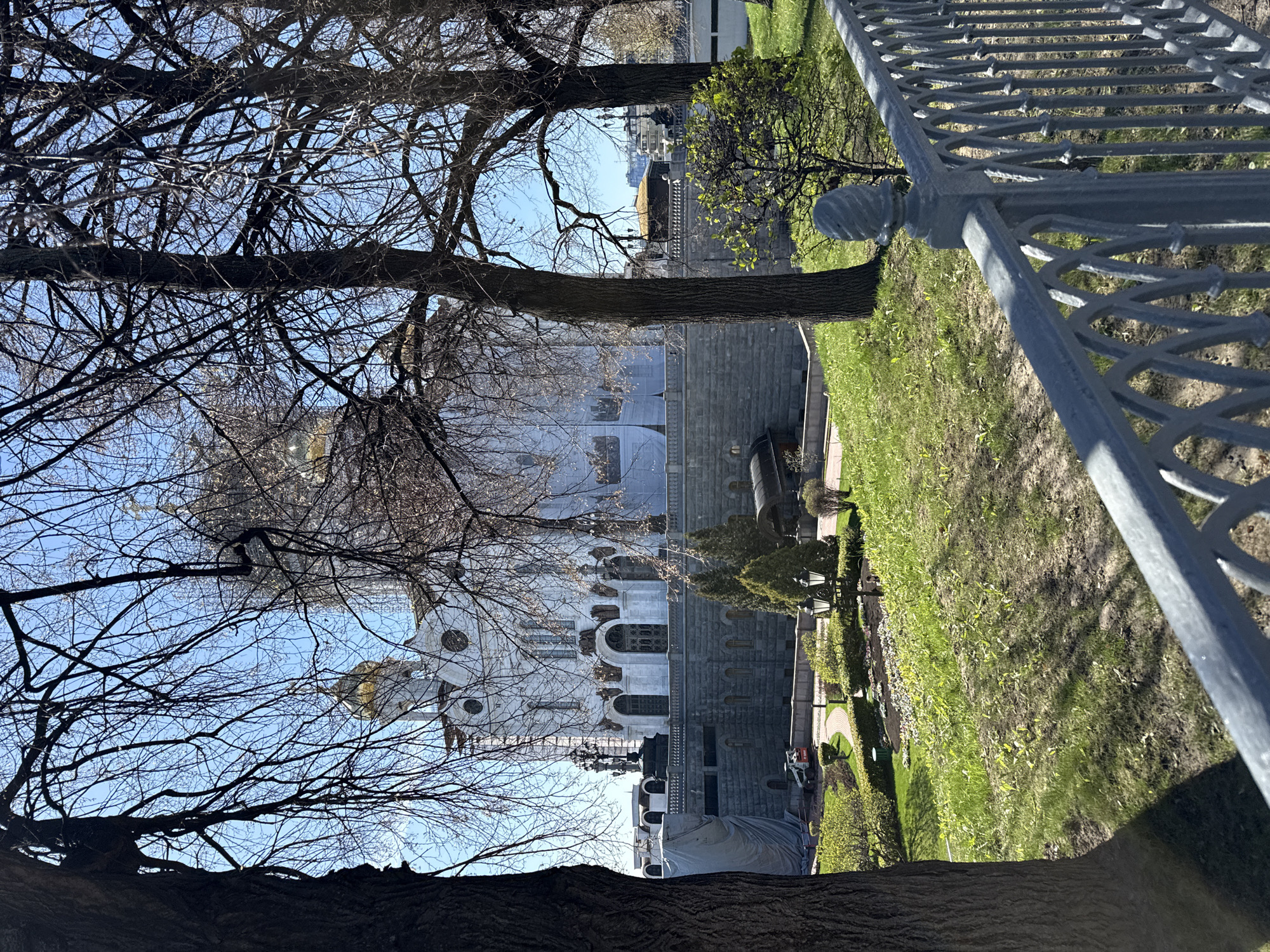 This image captures a grand architectural and natural scene on a bright, clear day, likely in late winter or early spring. The dominant subject is a magnificent white building, characteristic of a Russian Orthodox cathedral, featuring multiple golden domes, ornate arches, and intricate stone carvings. Given the provided context, this is likely the Cathedral of Christ the Saviour in Moscow. The building is constructed with a combination of light-colored stone on its upper sections and a darker, rougher stone for its foundation.
In the foreground and framing the central structure, several large deciduous trees stand with their intricate, bare branches silhouetted against a brilliant blue sky, indicating the season. Lush green grass covers the ground, suggesting early spring growth, though some areas appear patchy or shadowed. To the right, a decorative grey metal fence with distinctive circular and crisscross patterns provides a visual boundary.
Within the cathedral's landscaped grounds, smaller green bushes with emerging leaves are visible, alongside what appear to be pathways and old-fashioned streetlights. A large object, possibly covered for protection or maintenance, is situated near the base of the white building. Additionally, a cannon-like object is positioned on the grassy area, adding to the historical ambiance. The bright sunlight casts strong shadows, suggesting it is either morning or afternoon. No people or specific activities are discernable, contributing to a serene and majestic atmosphere. There is no visible text in the image.