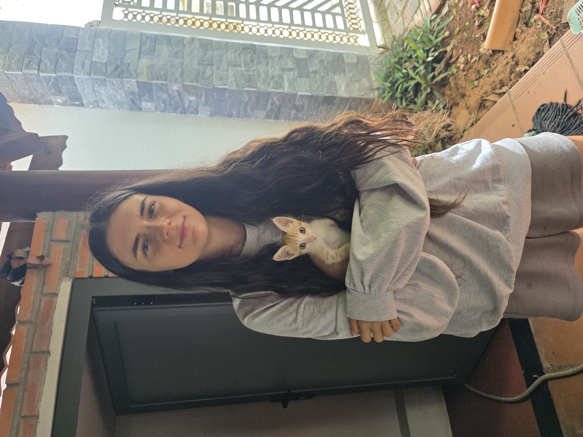 A young woman with long, dark, wavy hair is captured in a vertical, waist-up shot, holding a small kitten. She is wearing a light grey long-sleeved top and a matching skirt or dress. She looks directly at the camera with a gentle smile. Cradled in her arms, nestled against her chest and shoulder, is an adorable orange and white kitten with bright, light-colored eyes, also gazing towards the viewer.
The setting appears to be an outdoor or semi-outdoor area, likely a patio or balcony, in Mũi Né, Vietnam. The background features a distinctive stacked stone wall in shades of grey and white, partially obscured by a decorative white metal railing with circular motifs. Below this, a plain white wall is visible, along with a dark wooden structural beam on the left. To the right, there's an earthy area with green foliage, suggesting a garden space. The ground is covered with terracotta-colored tiles. A black rectangular object, possibly a TV or monitor, is present towards the bottom left of the frame. The lighting is natural and bright, indicating daytime. The scene conveys a warm, affectionate, and relaxed mood between the woman and the kitten. No visible text is present in the image.