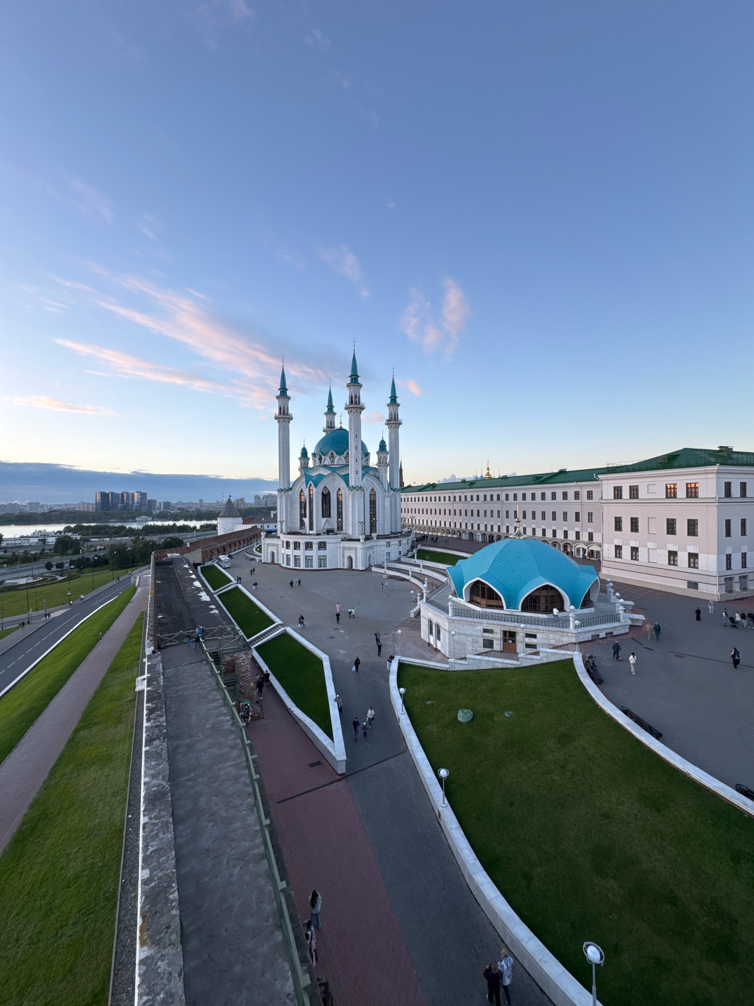 The image captures an elevated view of the Kul Sharif Mosque in Kazan, Russia, during what appears to be either sunrise or sunset, given the soft lighting and pastel colors in the sky. The mosque, a prominent landmark in Kazan, is characterized by its white facade, multiple minarets topped with turquoise spires, and a large turquoise dome. In front of the mosque, there is a distinctive blue-domed pavilion.
The surrounding area is a plaza, paved with stone and featuring manicured green spaces with sloping edges. Several people are visible walking around the plaza, suggesting a public space that is accessible. To the left of the mosque, a road with green verges runs alongside a walkway. In the background, across a body of water, the cityscape of Kazan is visible, with modern buildings and a hazy horizon. The overall scene is one of architectural grandeur and a serene urban environment.