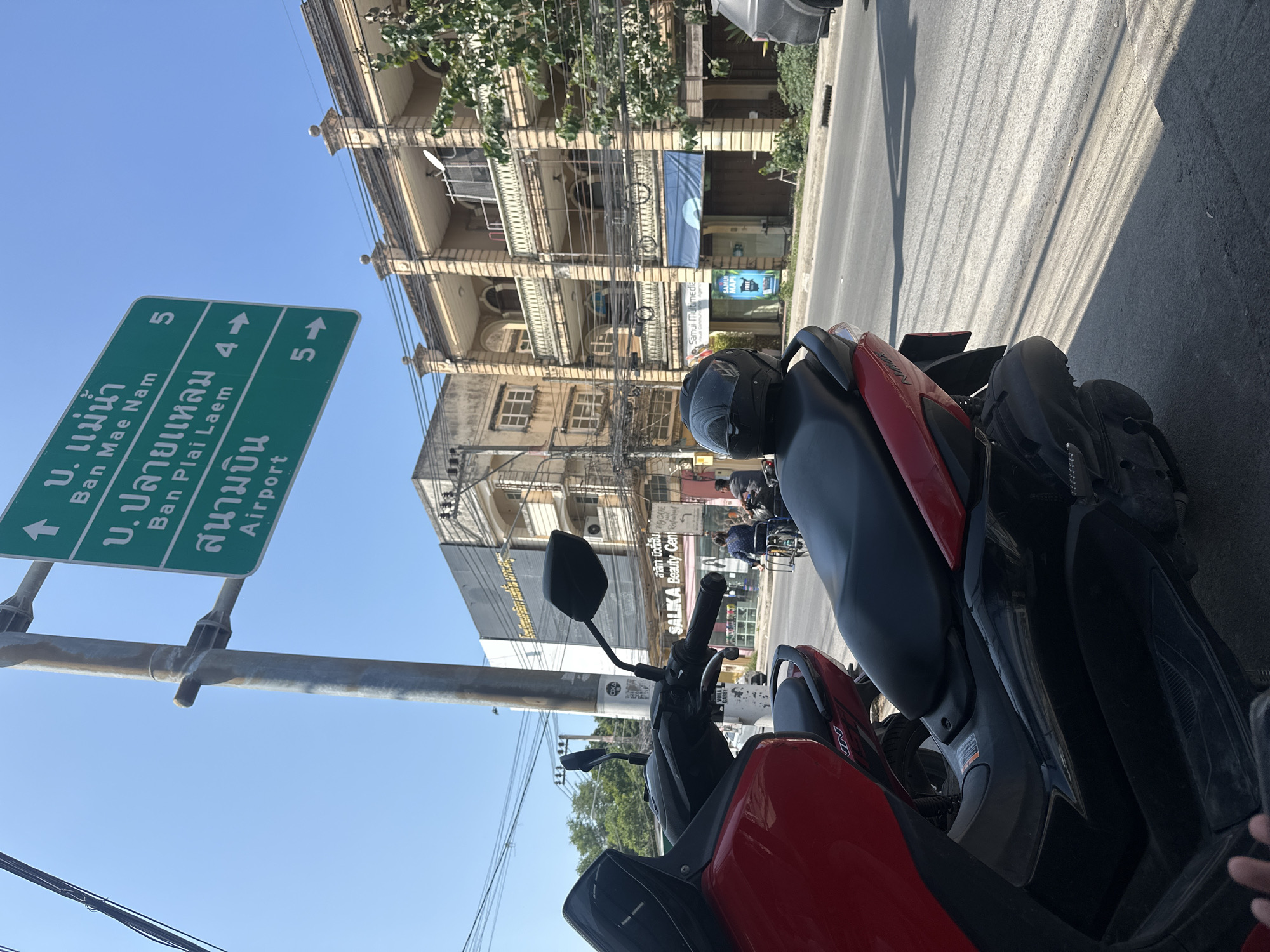 A sideways, eye-level outdoor shot shows a red and black scooter parked near a green road sign with white text, likely in Baan Bo Phut, Thailand. The sign indicates directions and distances to "Ban Mae Nam" (5 km), "Ban Plai Laem" (4 km), and the airport (5 km). In the background, two multi-story buildings with ornate facades are visible. A power line is strung across the buildings. Street view behind the scooter shows shops, some people, and another vehicle. The street surface is visible on the right.