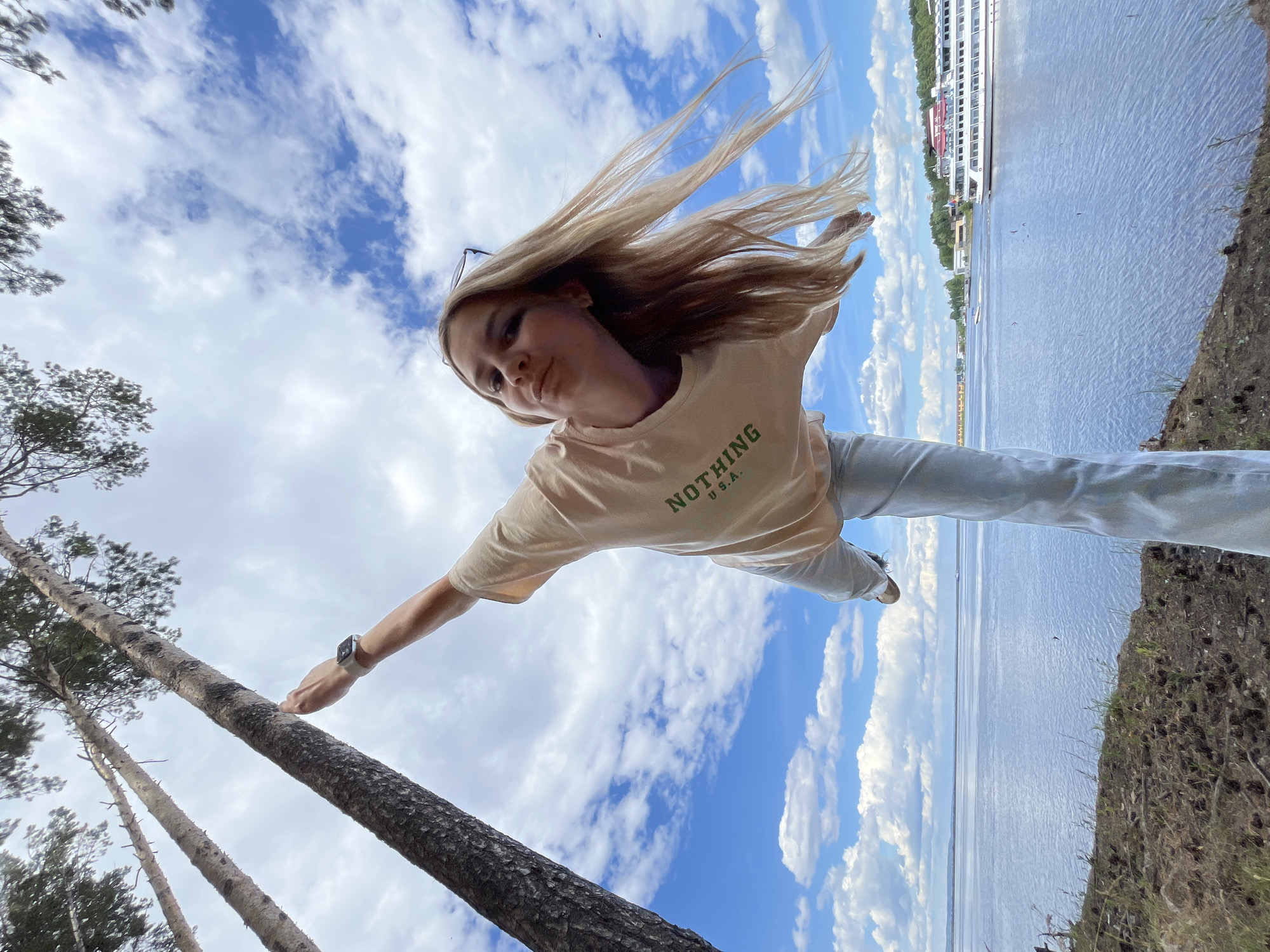 A young woman is captured in a dynamic, upside-down pose, with her body extending horizontally against a backdrop of a bright blue sky filled with fluffy white clouds. Her long blonde hair is windswept, adding to the sense of movement. She is wearing a light-colored t-shirt with green text that reads "NOTHING U.S.A." and light-colored pants. A smartwatch is visible on her left wrist.
Her left arm is outstretched, and her hand is touching a tall pine tree trunk, which is angled diagonally in the frame, drawing the viewer's eye from the bottom left upwards. Her right arm is extended behind her. The woman's face is turned towards the camera, and she has a playful expression.
In the background, to the right of the frame, a wide body of water stretches out, reflecting the sky. A large white ship with multiple decks is docked on the far bank of the water, with buildings and trees visible behind it. The scene suggests an outdoor setting, possibly a riverbank or a high vantage point overlooking a waterway. The bright sky and dynamic pose indicate it's likely daytime. The overall impression is one of freedom and exuberance.