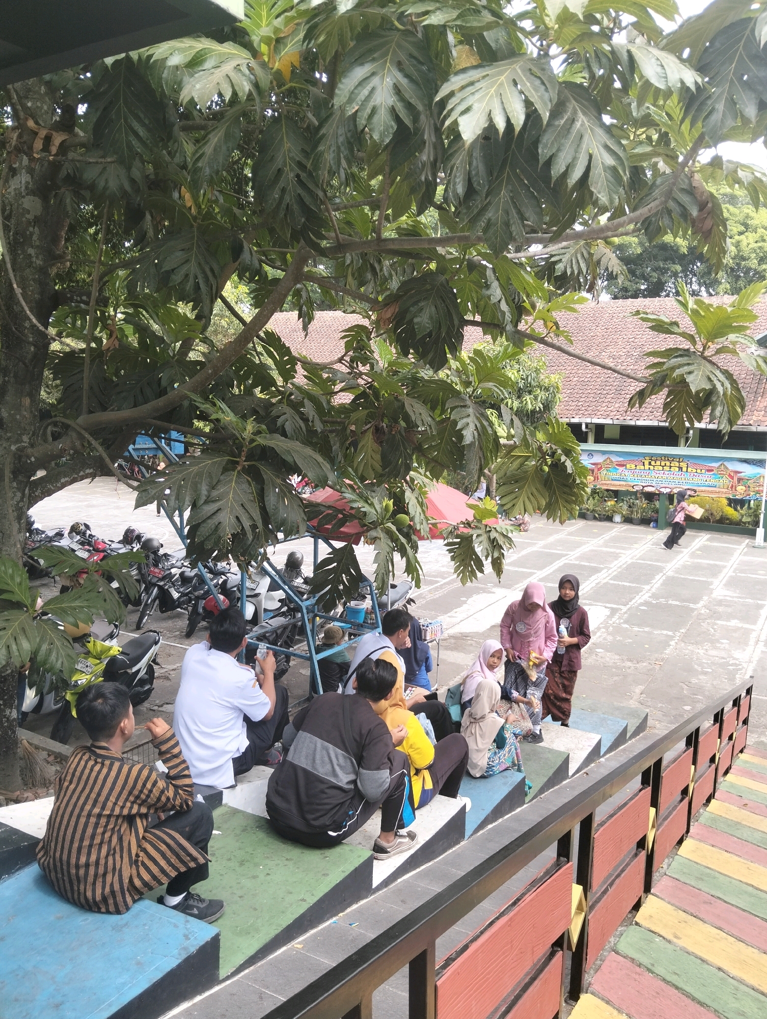 In Magelang, Indonesia, a group of individuals, some in traditional attire, are gathered outdoors on a tiered seating area under the shade of a large tree. The scene appears to be at a school or community event, indicated by a banner in the background with text that includes "FESTIVAL". Several motorcycles are parked in the background, suggesting a mode of transportation for attendees. The individuals are casually dressed, with some wearing hijabs. One person is seated on the steps, looking at their phone, while others are engaged in conversation. The tiered seating is painted in various colors, including blue, green, and yellow, with a colorful ramp leading to the side. The overall atmosphere seems relaxed and communal.