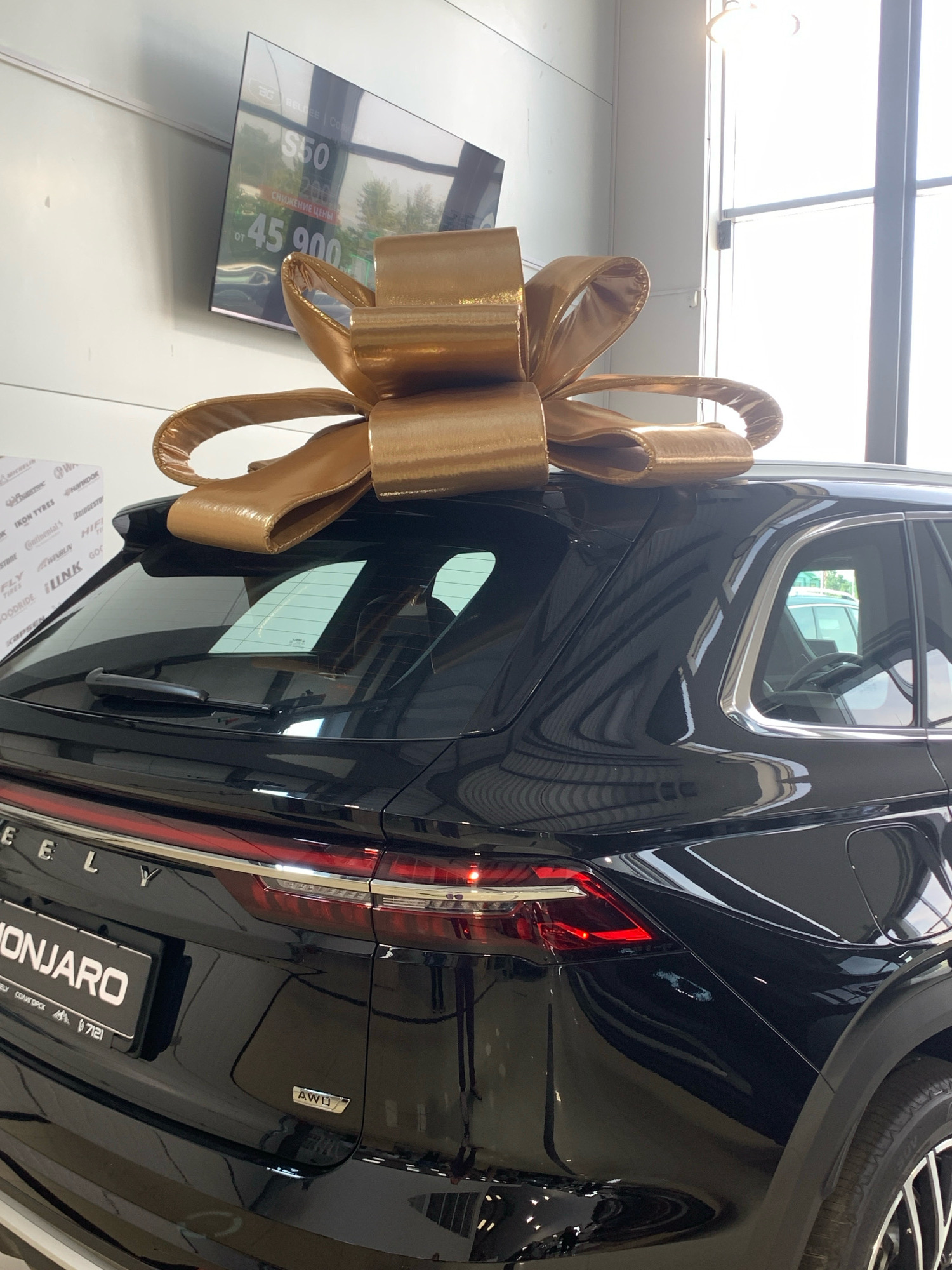 A black Geely Monjaro SUV is parked indoors, likely in a car dealership, with a large gold bow affixed to its roof. The vehicle's rear is visible, featuring its taillights, "GEELY" and "MONJARO" badging, and an "AWD" emblem. Reflections on the car's glossy surface indicate an indoor environment with bright lighting. In the background, a large screen displays pricing information, with "ОТ 45 900" prominently visible. The context of the "Чыжэвіцкі сельскі Савет" suggests the location is likely within or near this rural administrative unit in Belarus, possibly at a dealership in a town or village within its jurisdiction. There are no people or animals visible in the image.