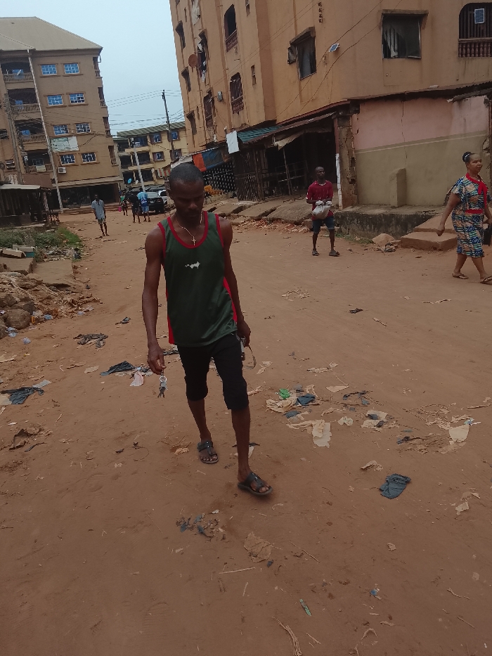 A dirt road in a neighborhood with tall buildings is littered with trash. In the foreground, a man in a tank top, capri pants, and sandals walks towards the camera, holding sunglasses and keys. Behind him, another man in a red shirt holds a bag. A woman in a patterned dress walks away on the right. Additional people are visible further down the road, and multiple buildings line the street.
