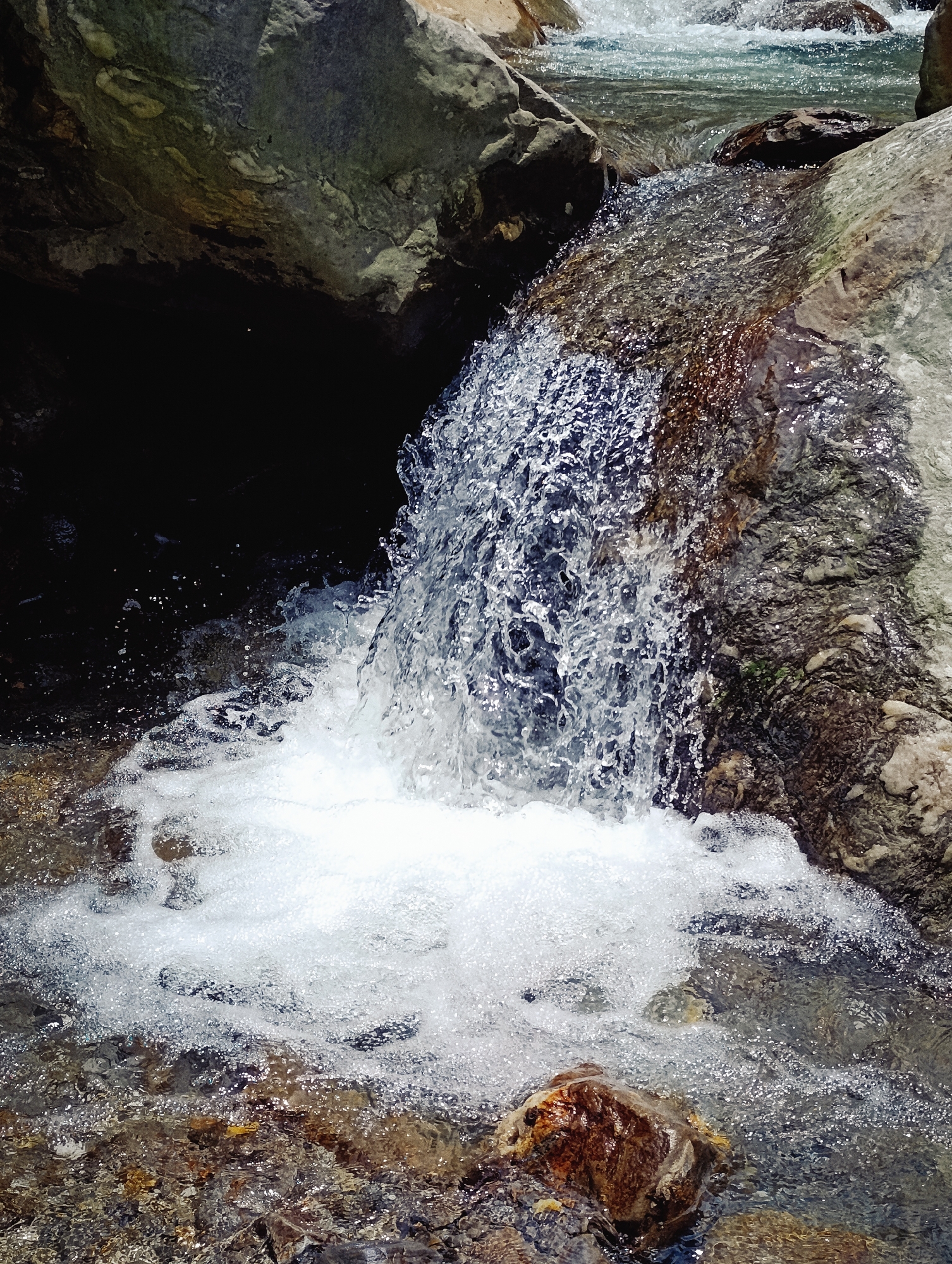 The image depicts a close-up view of a small waterfall cascading over rocks in a natural environment, possibly a riverbed. The water is clear and turbulent, creating white foam and spray as it hits the rocks below. The rocks are varied in color and texture, ranging from smooth, light-colored stones to rougher, darker ones. Some rocks appear to be partially submerged in the water. The background includes darker, larger rock formations, one of which forms a dark recess behind the waterfall. Sunlight glints off the water's surface, highlighting the movement and texture of the flowing water and the wet surfaces of the rocks. The scene suggests a dynamic and refreshing natural setting.