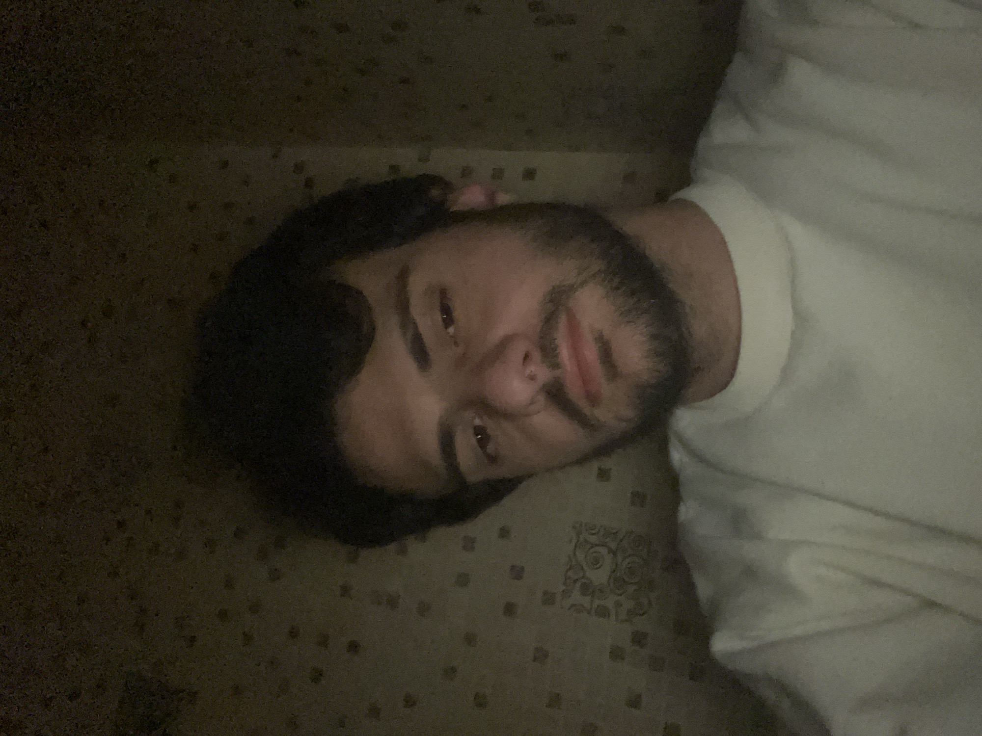 A man with dark hair and a beard is captured in a close-up, horizontal portrait, lying down indoors. He wears a light-colored, likely white, t-shirt. His head is tilted, and he looks directly into the camera with a neutral to slightly smiling expression. The lighting in the room is dim, suggesting an evening or nighttime setting. He is resting on a patterned surface, possibly a carpet or a bedspread with repetitive geometric motifs. The man's right eye shows a slight reddish tint, likely due to lighting conditions or reflection. No text is visible in the image. The scene takes place in Tbilisi, Georgia.