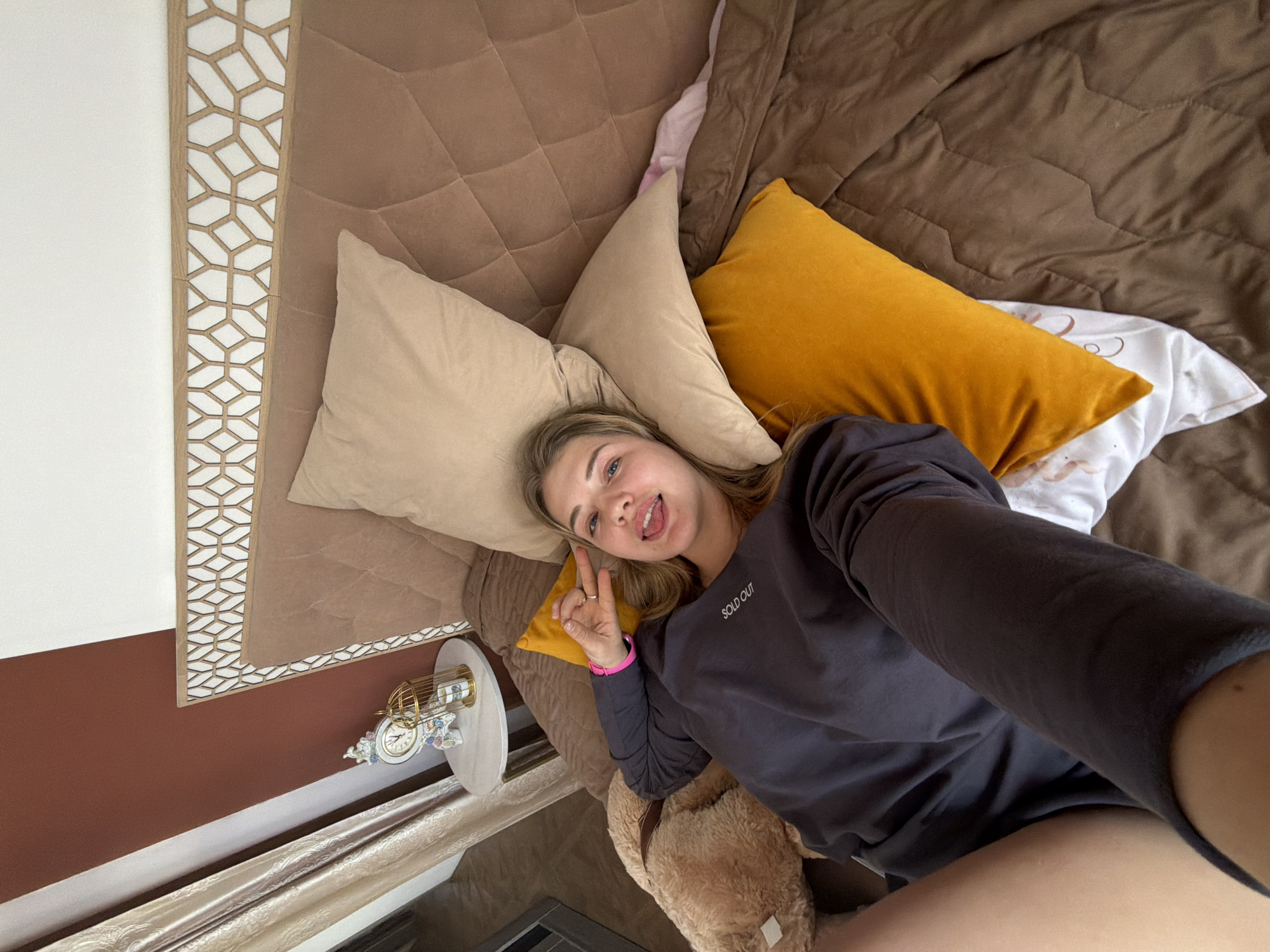 A young woman is lying on a bed taking a selfie. She is surrounded by pillows in shades of brown and yellow. The bed has a brown headboard with a geometric design on the sides. The woman is wearing a dark gray long-sleeved shirt. She is making a peace sign with her fingers and has braces on her teeth. A teddy bear is visible near her. A bedside table with a clock and other decorative items is next to the bed. The wall behind the bed is painted brown and white. The location is indicated as Большая Шильна, Russia.