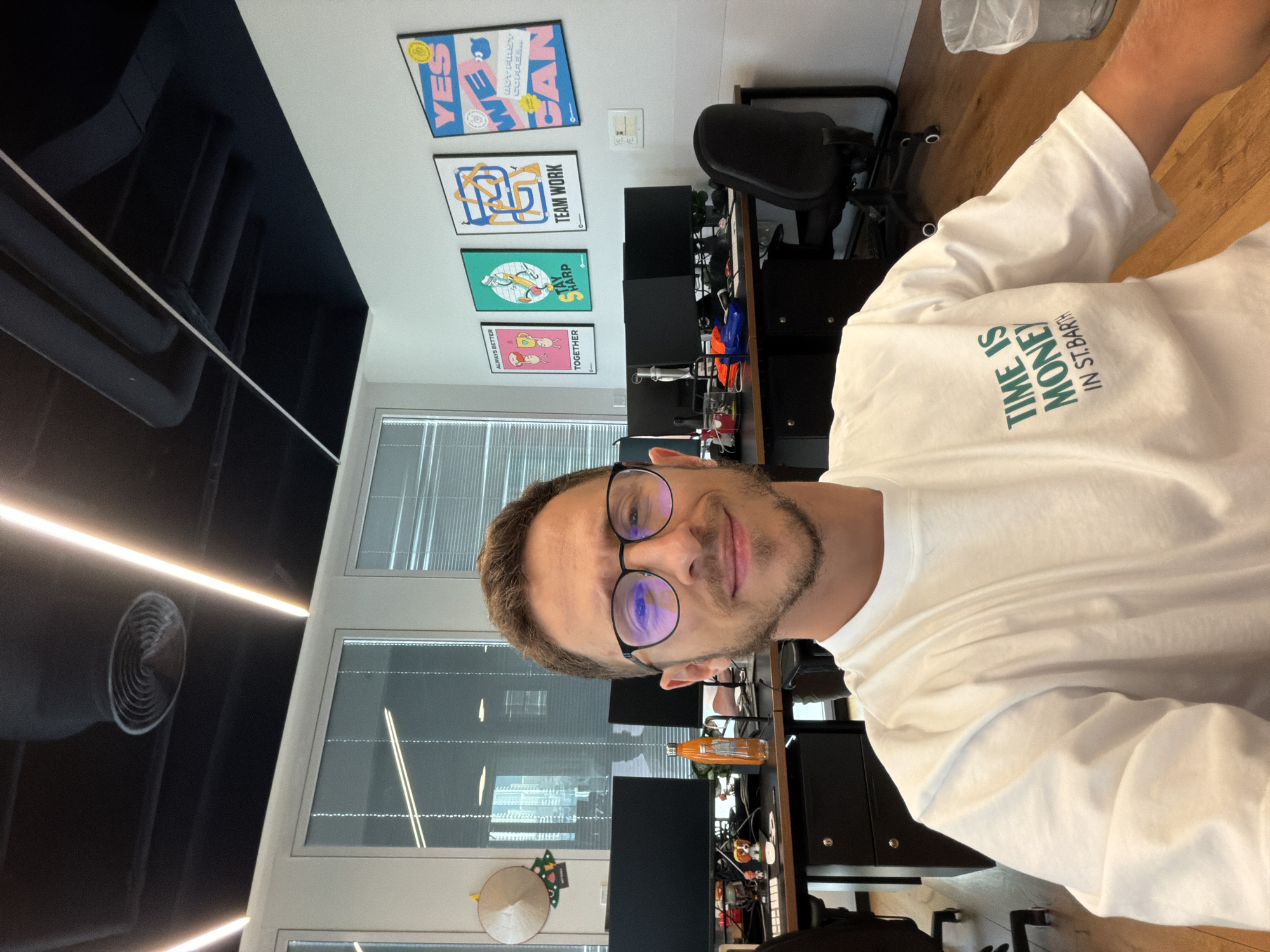 A man in a white t-shirt with the words "TIME IS MONEY IN ST. BARTH" is standing in an office setting. Behind him is a wall with posters and a window with blinds. Desks with computer monitors and chairs are also visible. The location may be Sdot Negev Regional Council, Israel.