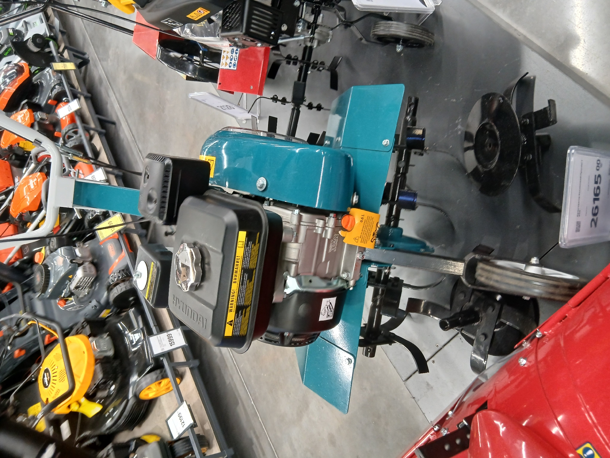 The image shows a close-up view of several tillers and other gardening equipment displayed in what appears to be a retail store. In the foreground, a teal-colored tiller with a black engine is prominently featured. It has a metal casing with yellow warning labels and a silver fuel cap. The tiller has rotating blades at the rear and a metal guard. To the left, the edge of a red piece of equipment, possibly another tiller or a wheelbarrow, is visible. In the background, more gardening tools, including orange tillers and black lawnmowers, are arranged on shelves. The flooring is concrete. A price tag with the number "26165 00" is visible at the bottom right corner. Based on the presence of multiple tillers and other gardening equipment, this scene is likely located in a hardware store or a specialized garden equipment shop. Given the mention of Chabany, Ukraine, it is plausible that this establishment is situated in or near that city, serving as a retail point for such items.