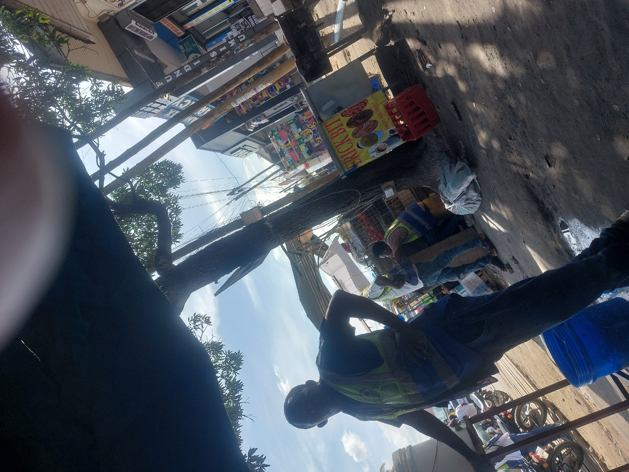 The image, possibly taken in Dar es Salaam, Tanzania, shows an outdoor urban scene. A man wearing a blue vest and jeans is in the foreground. In the background, there are several people, possibly workers, and a partially visible food stall labeled "ERICK BIT". Above the stall are commercial storefronts. The sky is visible with scattered clouds. The image is taken from a low angle and is slightly rotated. Trees and overhead wires are also visible.