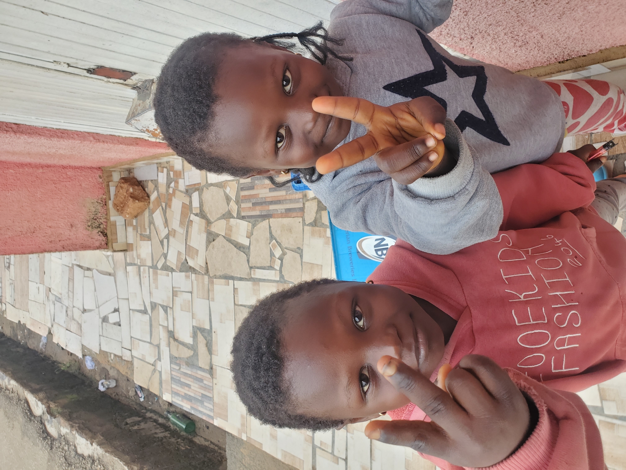 Two young children of African descent are standing on an outdoor walkway composed of irregularly shaped, beige and brown tiles. The child on the left, wearing a pink hooded sweatshirt with white lettering that reads "OOEKIDS FASHION 2046", is holding up two fingers in a peace sign. The child on the right, wearing a grey hooded sweatshirt with a black star design, is also holding up two fingers in a peace sign. Both children have dark skin and dark hair. The walkway is bordered by a drainage ditch on the left containing debris and a pink-colored wall on the right. The image appears to be taken from a slightly elevated angle looking down at the children. This scene is consistent with the general urban environment of Jos, Nigeria, characterized by paved walkways and open drainage systems.
