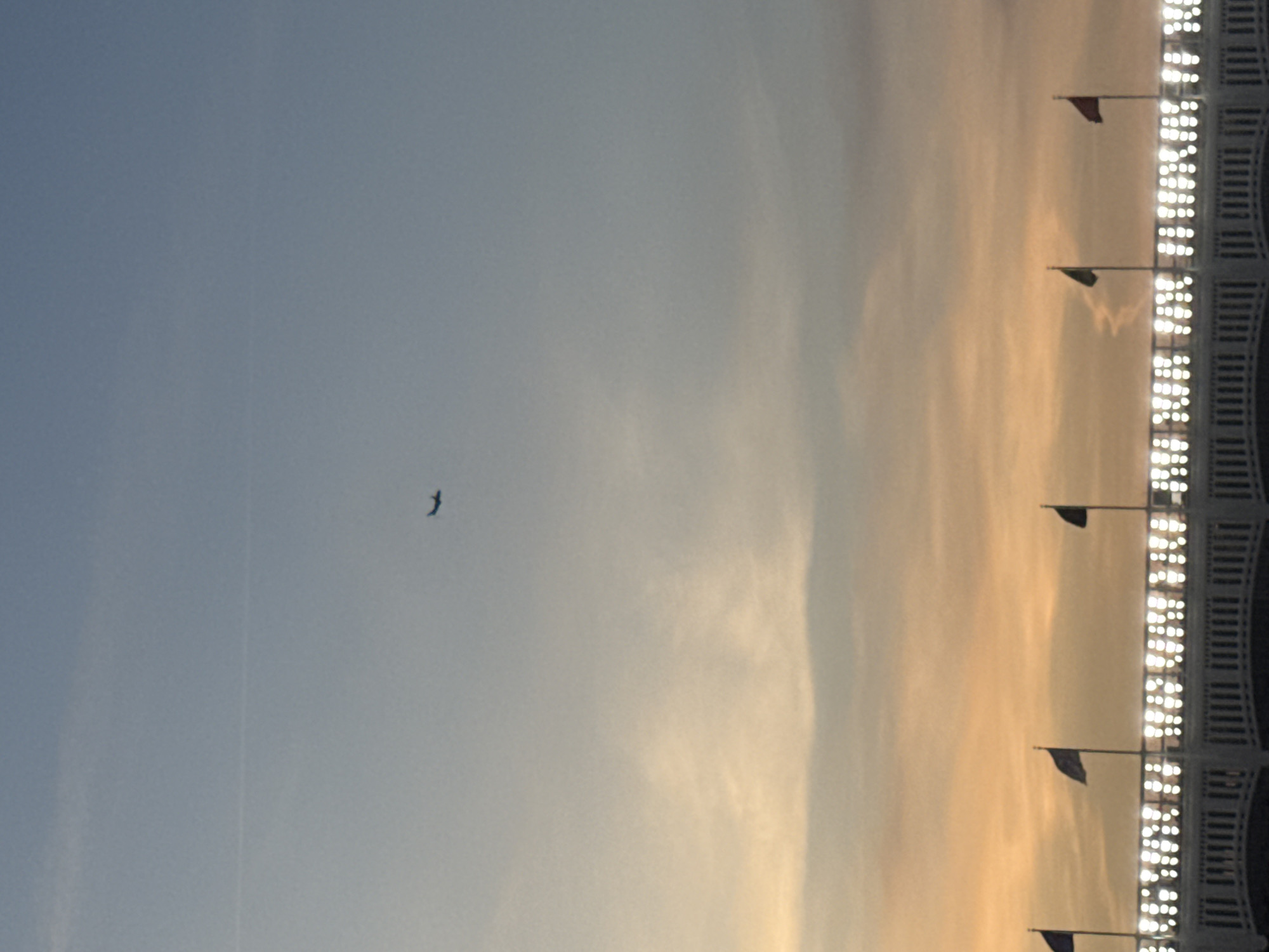 The image displays an exterior view of Yankee Stadium in the Bronx, New York City, against a twilight sky. The sky transitions from a light blue at the top to a warm orange-yellow closer to the horizon, indicating either sunrise or sunset. A faint contrail is visible in the upper-left quadrant of the sky. A dark silhouette of an airplane is captured in flight, positioned in the upper-mid-left portion of the frame. Below the sky, the distinctive white facade of Yankee Stadium is prominent. Five dark-silhouetted flags fly from poles atop the stadium structure. Below the flags, a line of numerous bright, circular lights illuminates the top edge of the stadium's arched architectural frieze. No people are visible in the scene.