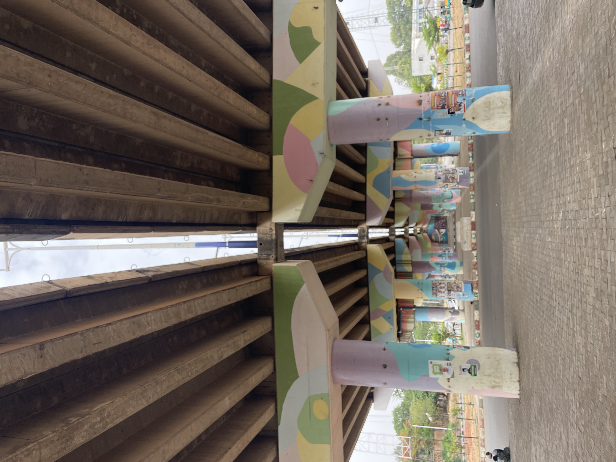 The image shows an underpass in Jos, Nigeria. The concrete structure features parallel beams overhead, supported by painted pillars. The pillars are decorated with geometric shapes in pastel colors and display various posters and stickers. The ground is paved with light-colored bricks or concrete blocks. In the background, trees and buildings are visible, indicating an urban environment. A few people can be seen in the distance near the edge of the paved area. The overall perspective is looking up and through the underpass.