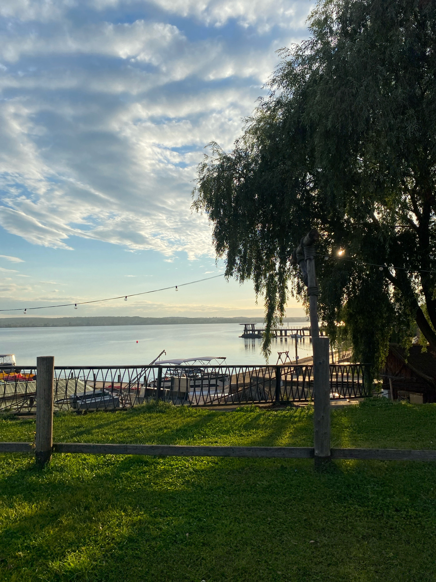 The image depicts a serene waterside scene, likely a park or recreational area, with a large weeping willow tree prominently featured on the right. The sky above is filled with scattered clouds, illuminated by the soft light of what appears to be late afternoon or early evening sun, which filters through the tree's branches. In the foreground, a well-maintained green lawn is visible, separated by a low wooden fence and a wooden post. Beyond the lawn, a black metal fence runs horizontally, behind which several boats are moored along a calm body of water. A distant shoreline with trees forms the horizon. The scene appears tranquil and peaceful, characteristic of a leisure spot. It is highly probable that this location is the Votkinsk Reservoir embankment in the city of Votkinsk, Russia, a popular spot for relaxation and recreation, known for its scenic views and green spaces.