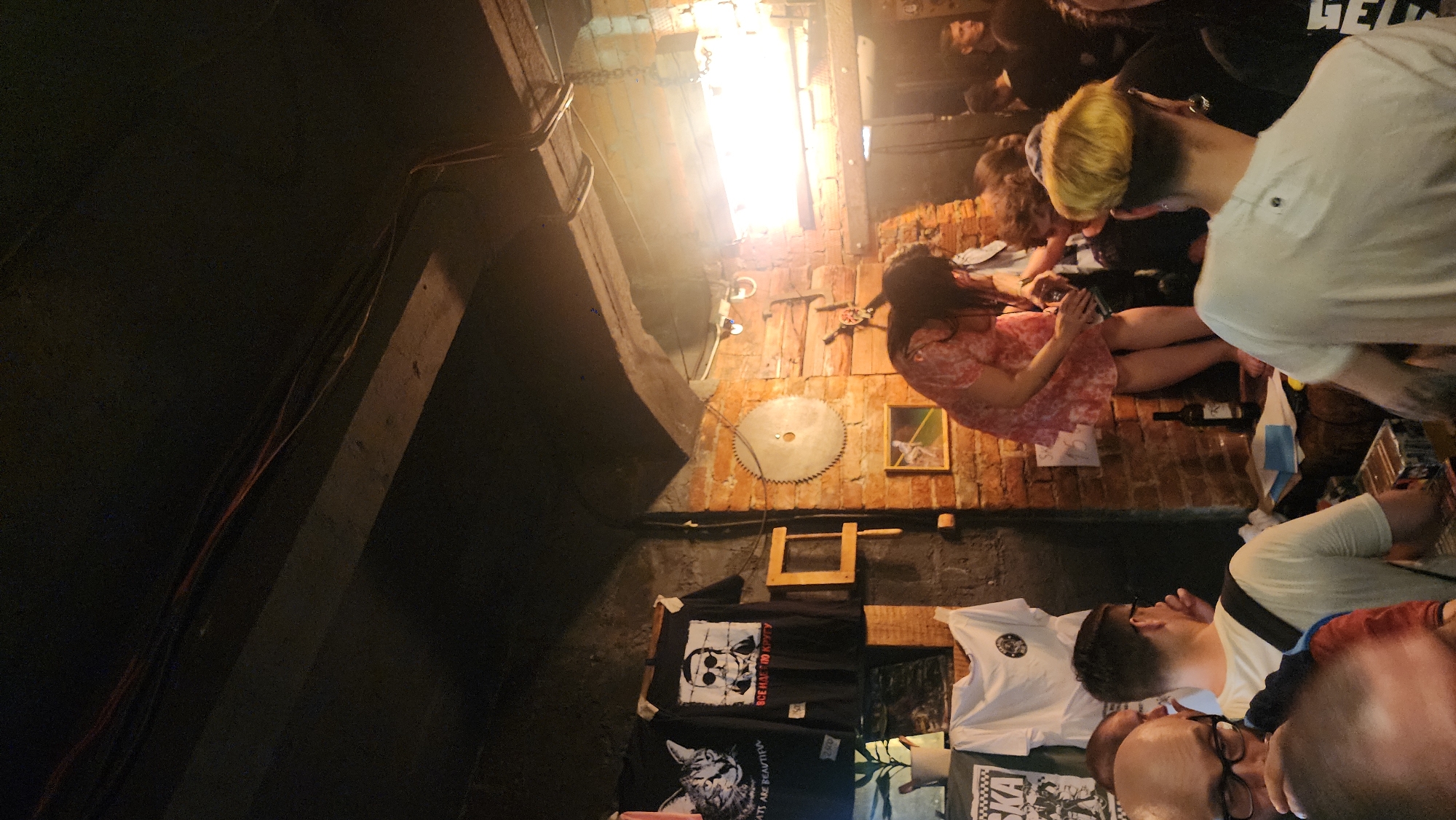 The image shows an indoor scene in Moscow, Russia. The walls are exposed brick, decorated with a circular saw blade, framed art, and wooden fixtures. Below these, t-shirts with various graphic designs are displayed. A group of people is present; one woman is seated on the floor wearing a pink patterned dress and looking at a mobile phone, near a bottle of wine. Other individuals are visible, some wearing white shirts, observing the scene. The space is dimly lit, with a light source above. There is a concrete structure to the left. The photograph is taken at an angle, making it difficult to ascertain precise dimensions.