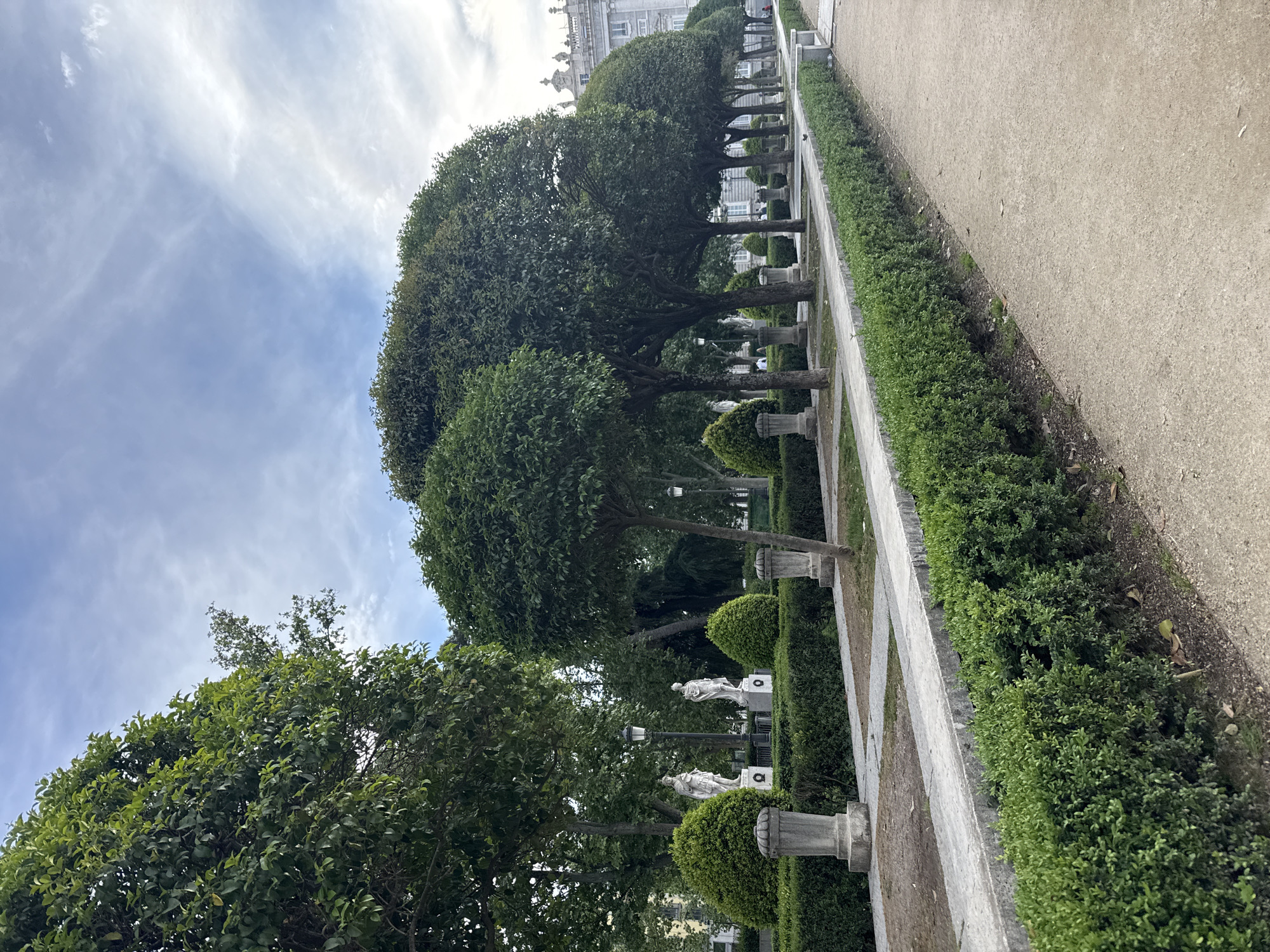 The image captures a formal garden setting in Madrid, Spain. A row of meticulously trimmed trees with round canopies lines the center of the frame, interspersed with classical stone urns and statues. A trimmed hedge borders the trees, separating them from a gravel path. In the background, architectural details of a building, likely a palace or government structure, can be discerned. The sky is partially cloudy.