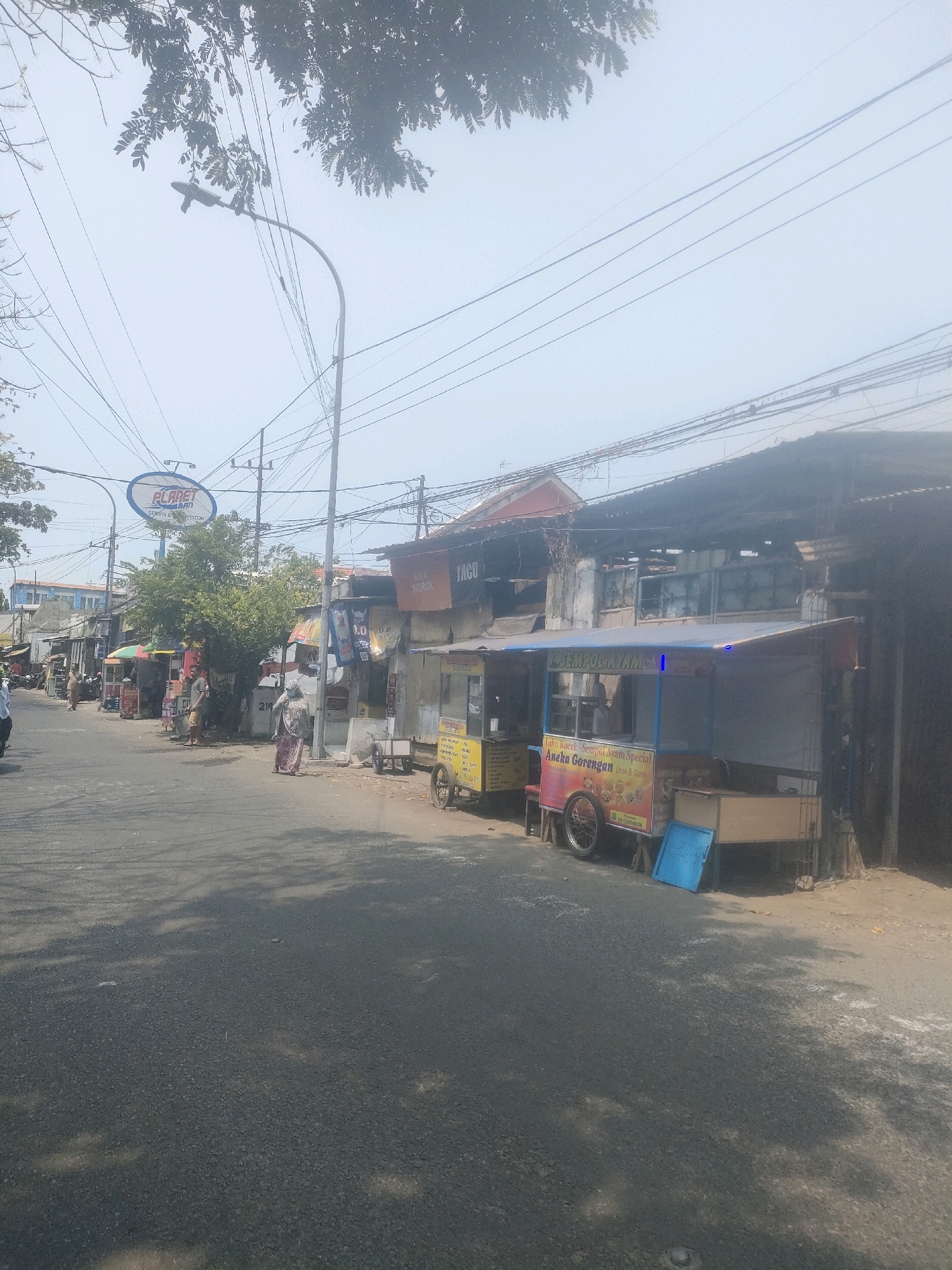 The image depicts a street scene in Surabaya, Indonesia, likely on a moderately overcast day. In the foreground, a paved road with shadows of trees stretches into the distance. To the right of the road, several street food vendors operate from carts. One cart, labeled "SEMPOL AYAM" and "TAHU KOREK - SEMPOL AYAM SPECIAL Aneka Gorengan Anak & Guru," displays food items and a phone number. Next to it, another vendor cart is visible. On the left side of the road, a row of shops and stalls lines the street, with some people walking and interacting. A prominent blue sign for "PLANET BAN" is visible above one of the shops, indicating a tire and auto service. Overhead, a complex network of electrical wires is strung between poles, common in urban Indonesian settings. The architecture suggests a mix of commercial and residential buildings, typical of a busy urban area.