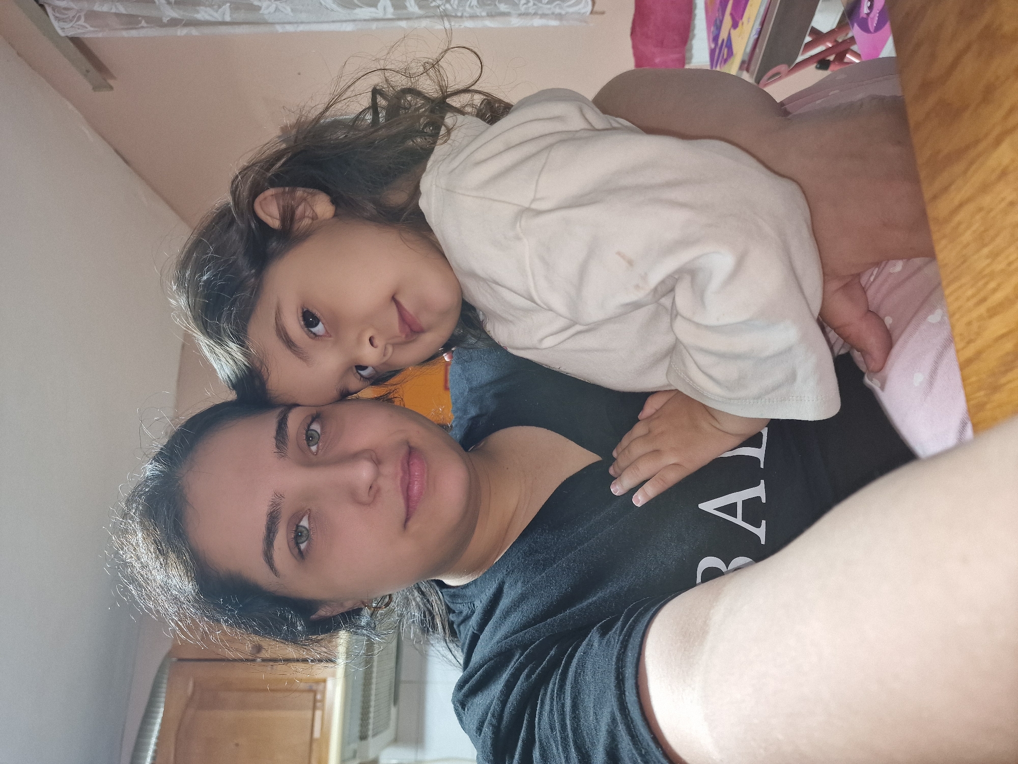 A selfie-style photograph shows a woman and a young child. The woman is wearing a black t-shirt with white lettering. The child is lying on top of the woman, wearing a white long-sleeved shirt. The background is out of focus, but kitchen cabinets and a window air conditioner can be seen. There is also what appears to be a child's chair or small desk. The location is in an unknown city in an unknown location.