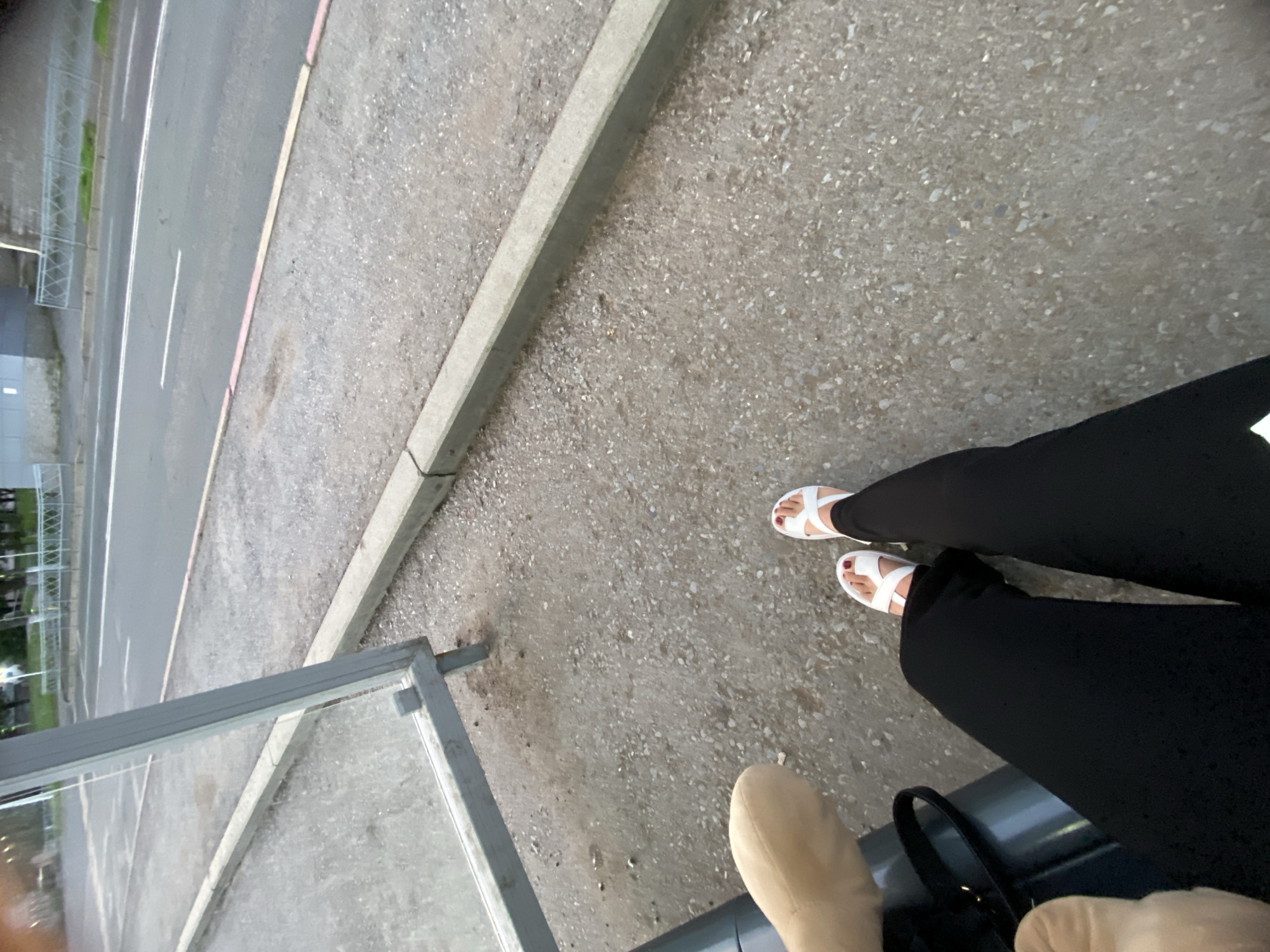 The image shows a person's legs and feet at what appears to be a bus stop. The person is wearing black pants and white sandals with red nail polish. In the background, a road with a bus stop shelter is visible. The ground is paved with gravel and concrete. The setting suggests an urban environment, possibly a bus stop in Ufa, Russia, given the prompt's request. There is also a hint of a tan-colored object, possibly a bag or another person's leg, to the left of the frame.