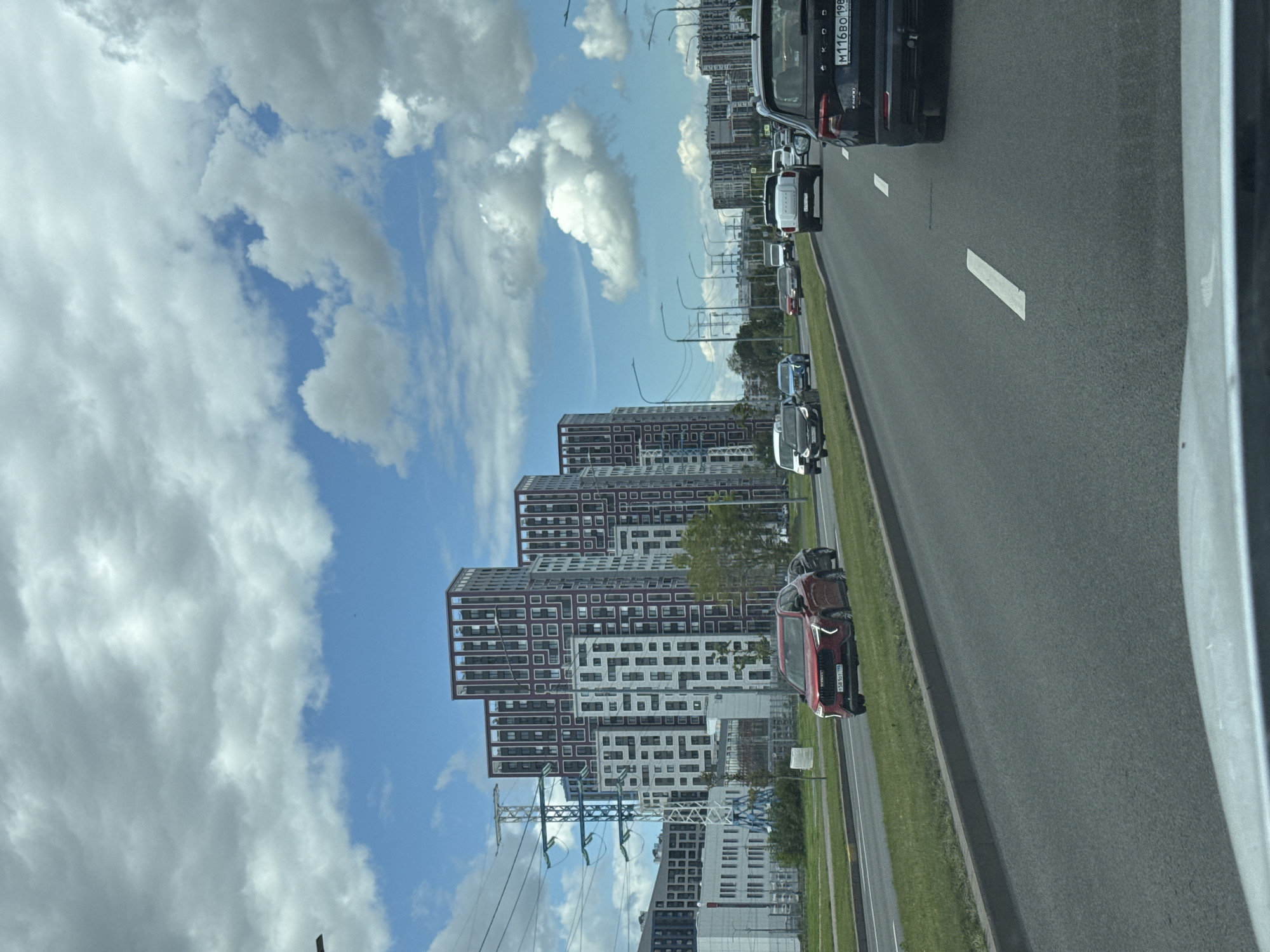 This image depicts a street scene in a modern city. In the background, a cluster of tall, contemporary buildings with a repeating pattern of windows and maroon accents dominates the skyline. To the left, a large electrical transmission tower with multiple crossbars and cables is visible, indicating urban infrastructure. The foreground shows a multi-lane asphalt road with visible white lane markings. Several cars are traveling in the same direction on the road. One red SUV is in the left lane, and a black SUV is in the right lane. The sky is partly cloudy, with large white cumulus clouds against a blue backdrop. The overall impression is that of a busy urban environment on a bright, possibly warm, day.