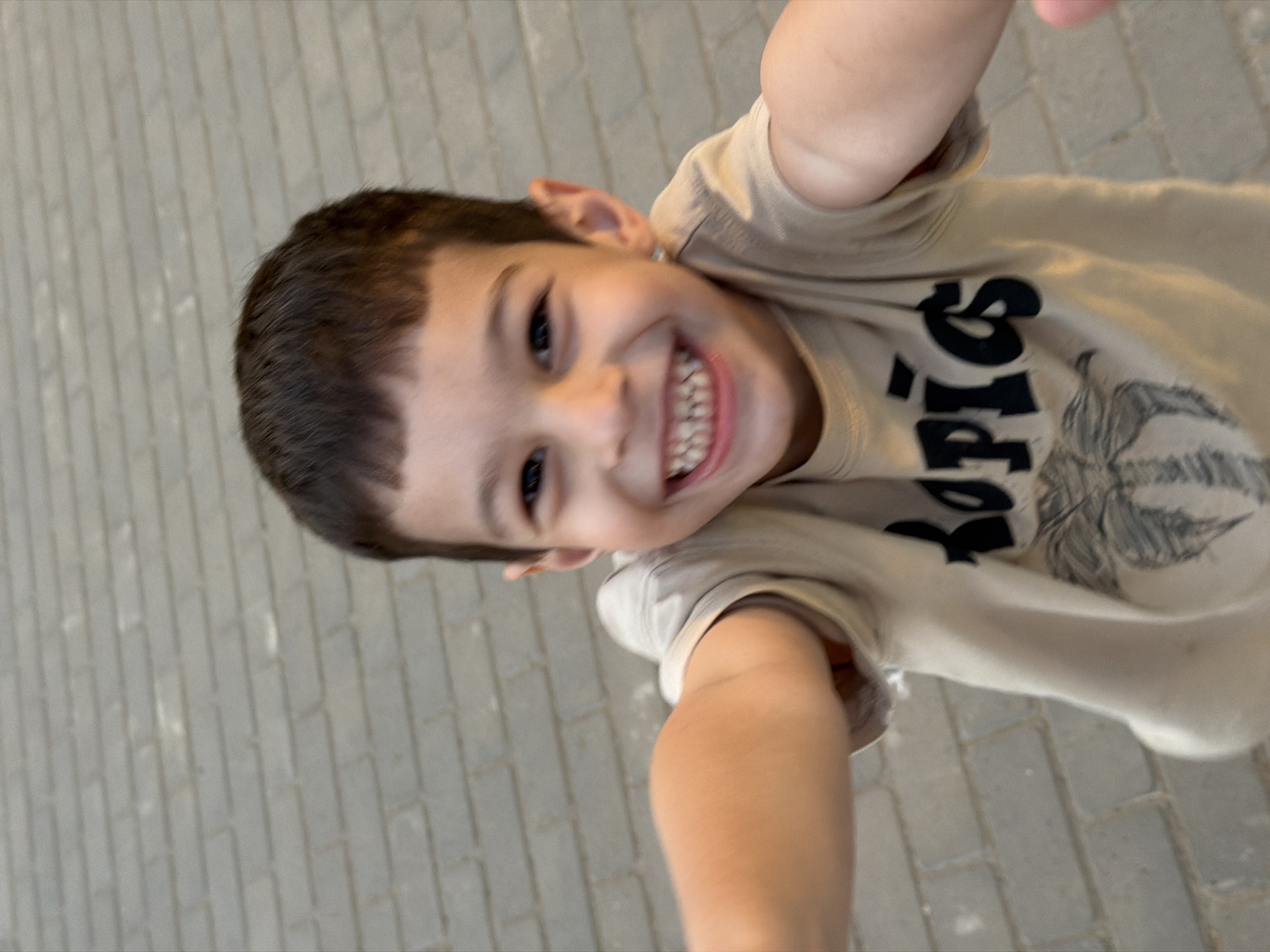 This is a close-up, slightly tilted, eye-level selfie of a young boy. He has dark hair, brown eyes, and is smiling widely, showing his teeth. He is wearing a light-colored t-shirt with black text that appears to read "PIGS" with a graphic of an animal's face on it. The boy's arm is visible in the foreground, and it appears he is holding the camera. The background is a light gray brick-like surface, suggesting an outdoor setting on a paved area. The lighting is bright, indicating it is daytime. The boy's expression is joyful and energetic. There are no other people or objects clearly visible in the frame.