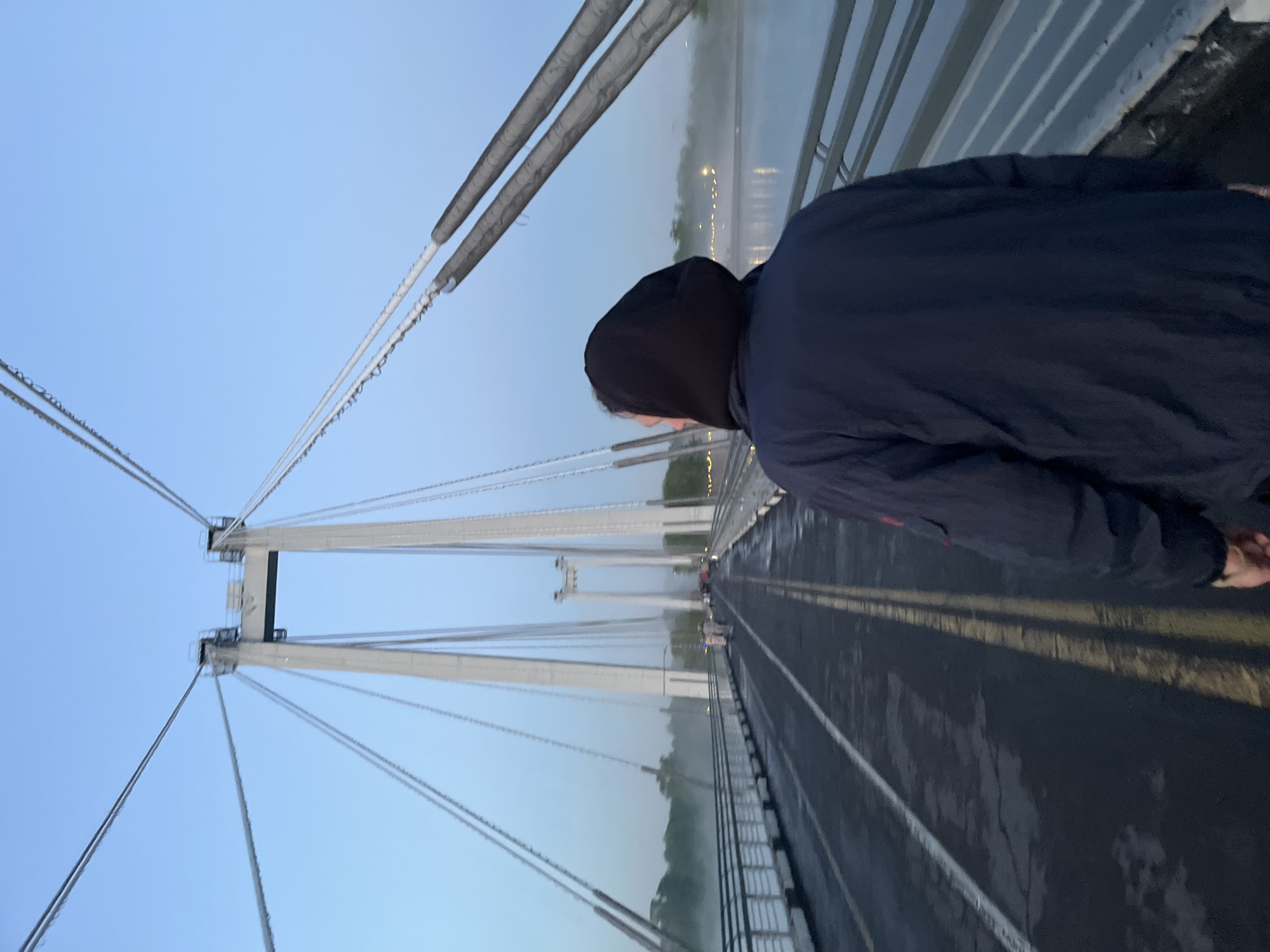 A person wearing a black hooded jacket stands on a bridge with prominent suspension cables and white support towers. The bridge appears to be wet, likely due to rain or mist. In the distance, there's a hazy view of what could be the city of Krasnoyarsk, Russia, as the bridge resembles the Vinogradovsky Bridge, a notable landmark in the city. The sky is a pale blue, indicating early morning or late evening light.