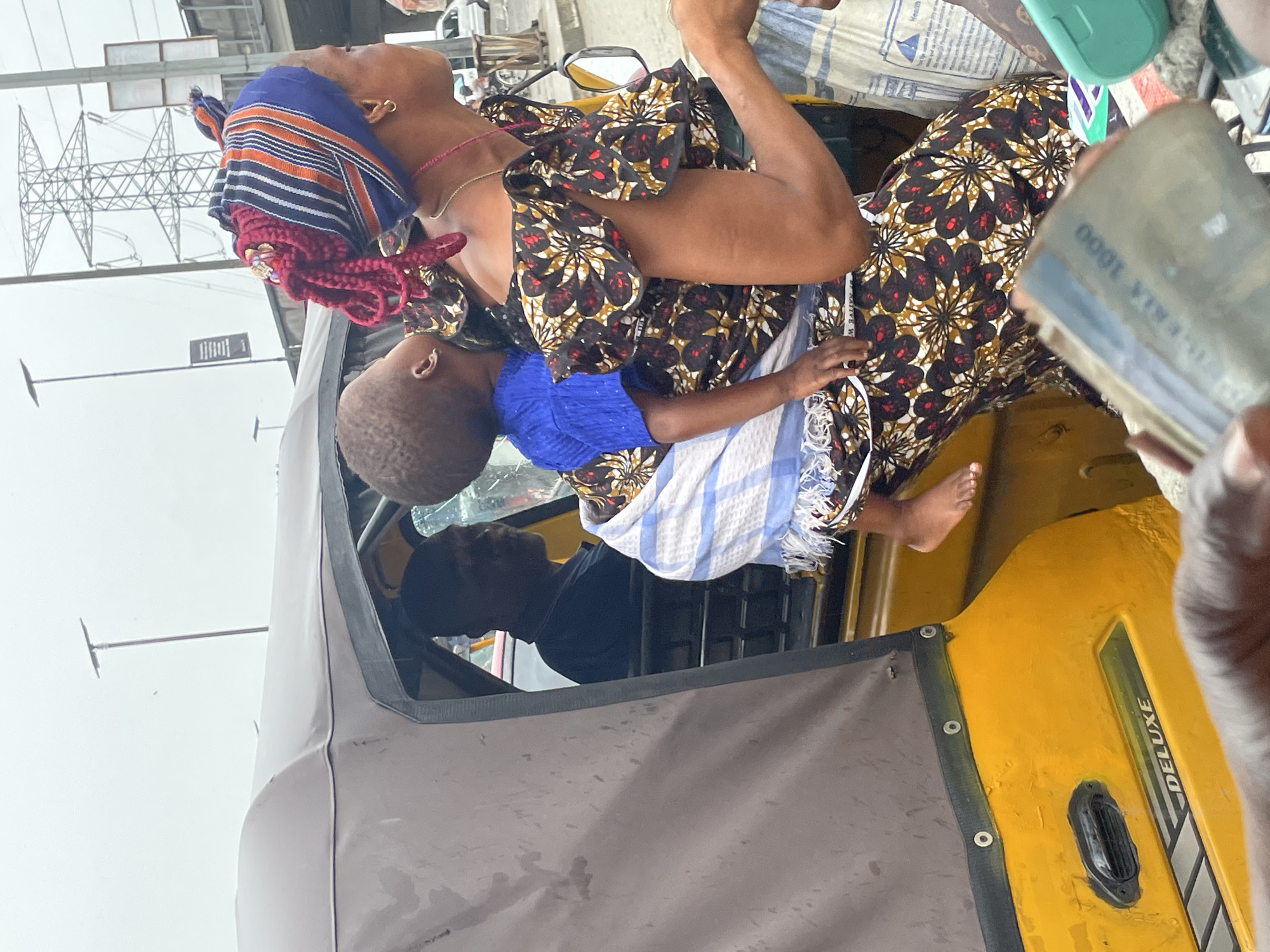 In Ado, Nigeria, the image shows a woman with a child in a yellow "Deluxe" branded auto-rickshaw (keke). The woman is wearing a patterned dress and a head wrap with red braids. The child is wearing a blue shirt and is wrapped in a blue and white striped cloth. The driver of the auto-rickshaw is visible. In the foreground, a hand holds out a 1000 Naira note. Overhead, power lines are visible. Other details: some bags, possibly containing food and other items, inside the vehicle.