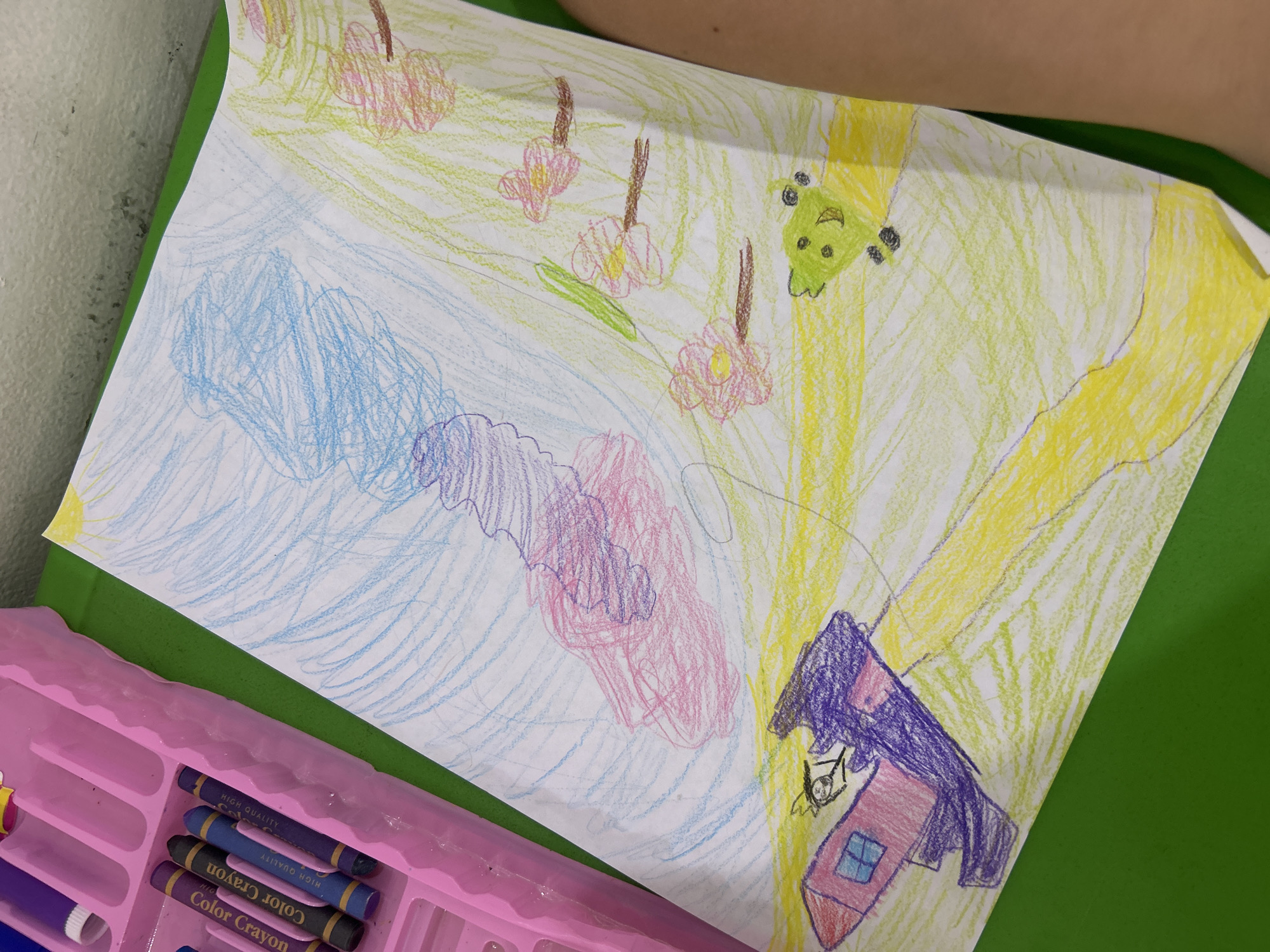 A child's drawing on white paper depicts a whimsical scene. In the sky, a large pink and blue cloud formation swirls, with a yellow sun in the upper right corner. Below, a purple and pink rocket ship with a small stick figure pilot ascends from a pink house with a blue window. A yellow path curves across the ground, which is colored in light green and yellow scribbles. Several pink flowers with brown stems are scattered across the scene, along with a green frog with black eyes. The drawing is placed on a green surface, with a pink crayon box visible in the upper left corner.