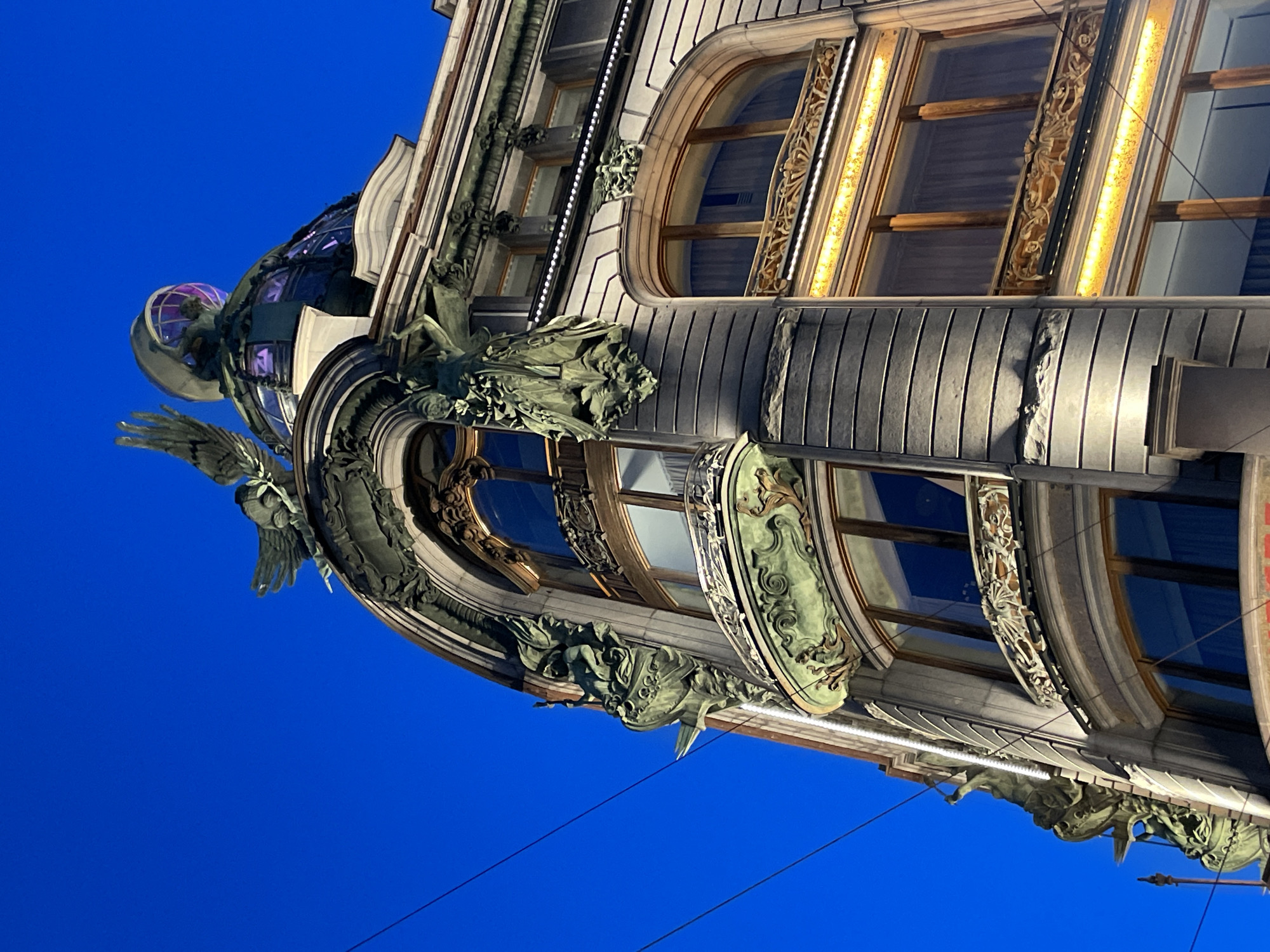This media file captures a highly ornate building facade viewed from a low angle, looking upwards towards a deep, clear blue sky. The building exhibits intricate architectural details, characteristic of Art Nouveau style.
The main subjects are the building's elaborate exterior and its numerous sculptures. At the apex of the building, a large, translucent glass globe or dome, emitting a soft purple light from within, is supported by a winged figure, possibly an angel or mythological creature. Further down, other sculptures in a muted greenish-bronze color, suggesting patinated metal, adorn the facade. These include additional winged figures, mythical beasts resembling dragons or sea creatures with detailed scales, and reclining human-like figures amidst decorative foliage.
The building features large, arched windows framed by richly detailed decorative elements, which are internally illuminated with a warm yellow glow, highlighting their ornate patterns. Sections of the facade are ribbed or paneled in a greyish material, contrasting with the decorative elements and sculptures.
The scene is set against a dark blue sky, strongly indicating evening or night, with the building's artificial lighting prominently visible. Thin black power lines are visible stretching across the lower part of the sky, suggesting an urban setting. The overall impression is one of grandeur and detailed craftsmanship.
No text is visible in the image. Given the context, the location is Saint Petersburg, Russia.