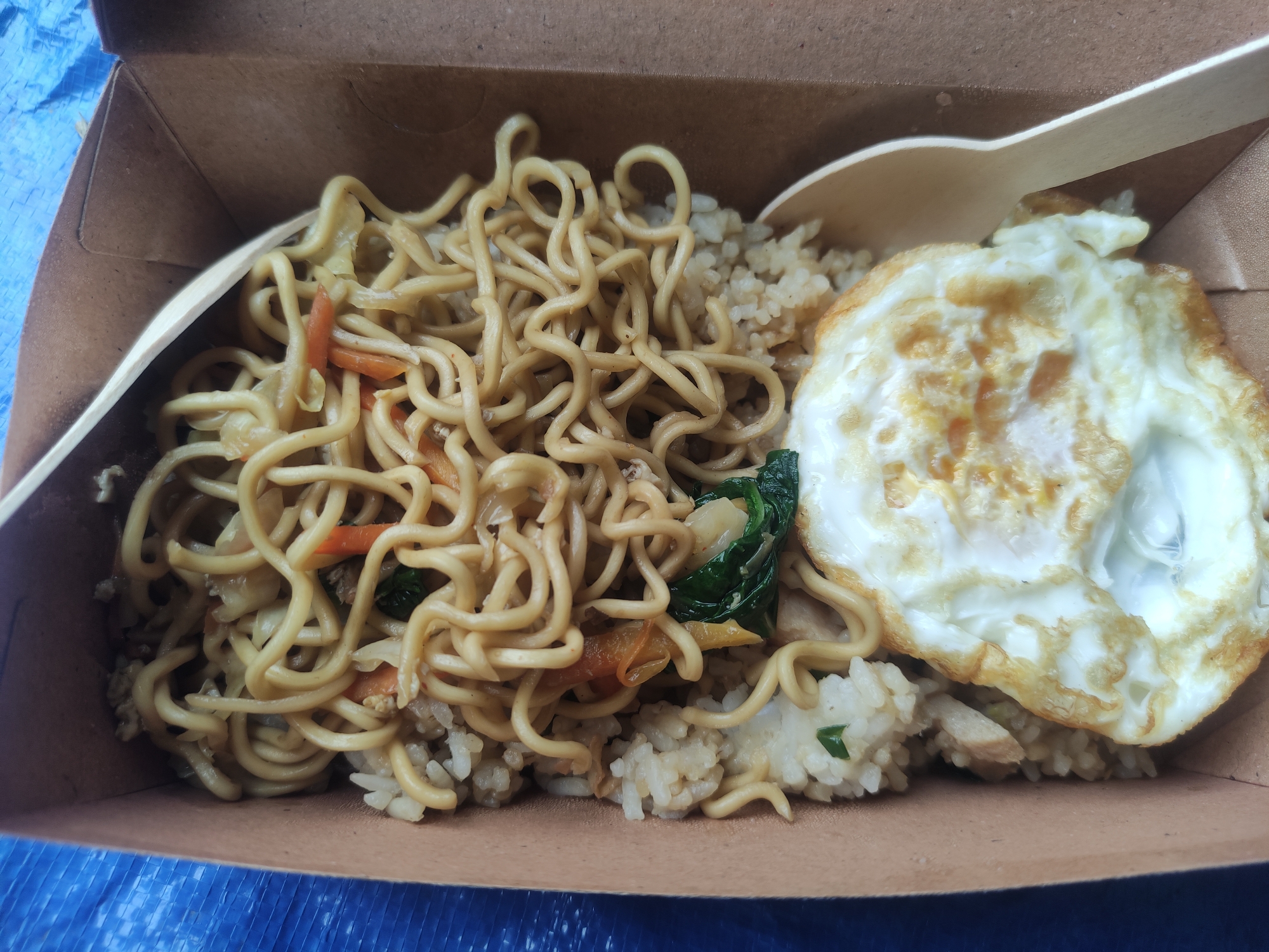 A cardboard container holds a serving of stir-fried noodles mixed with vegetables, positioned alongside a bed of rice and topped with a fried egg. A wooden spoon rests on the food. The meal is likely a takeout order. The image originates from Babakan Madang, Indonesia.