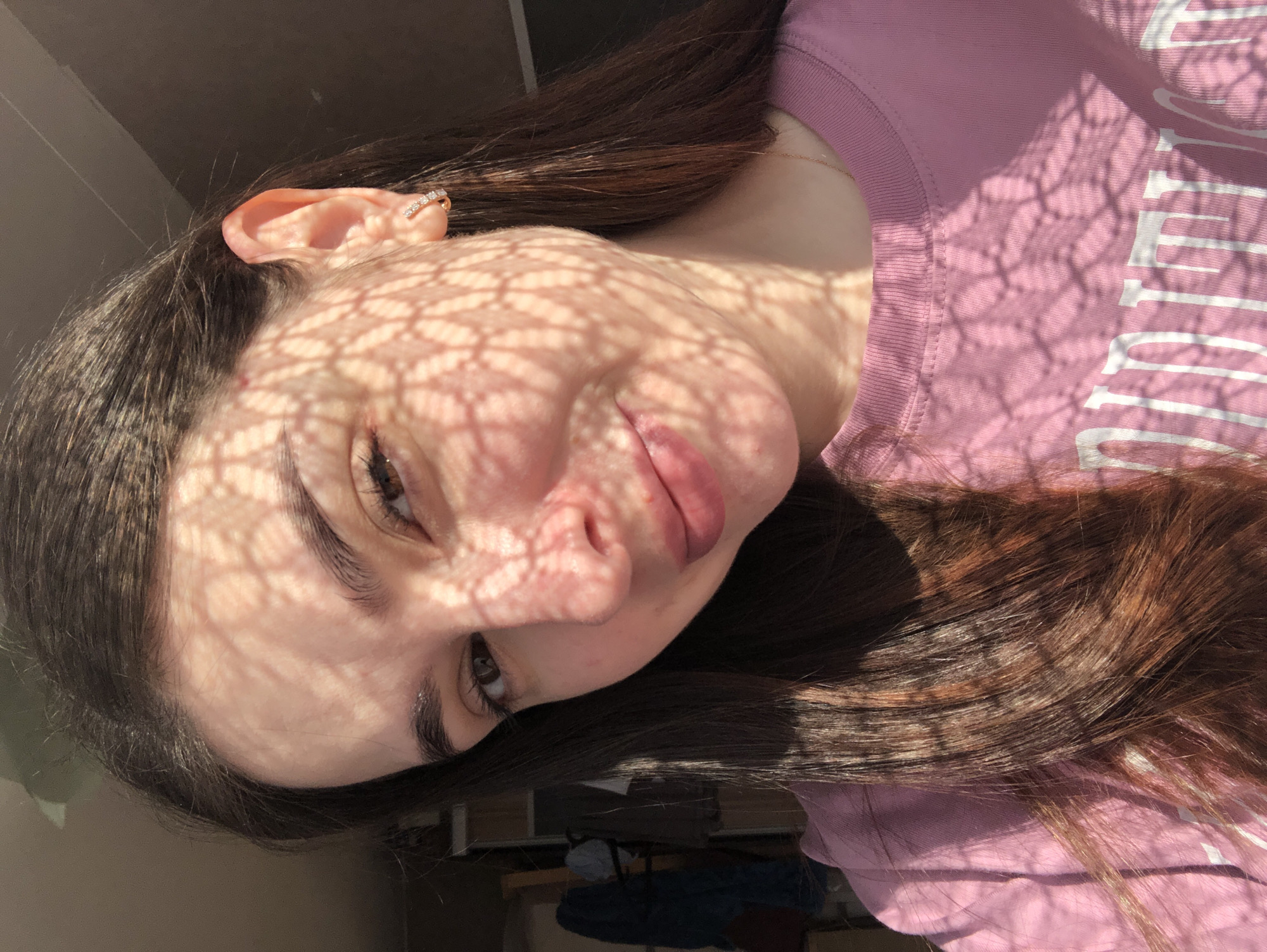 A young woman with dark brown hair is indoors. She is wearing a pink shirt and has an earring in her left ear. A shadow of a lattice-like pattern is cast across her face. The location is assumed to be Chaikovsky, Russia. A partially visible desk and blue object are present in the background.