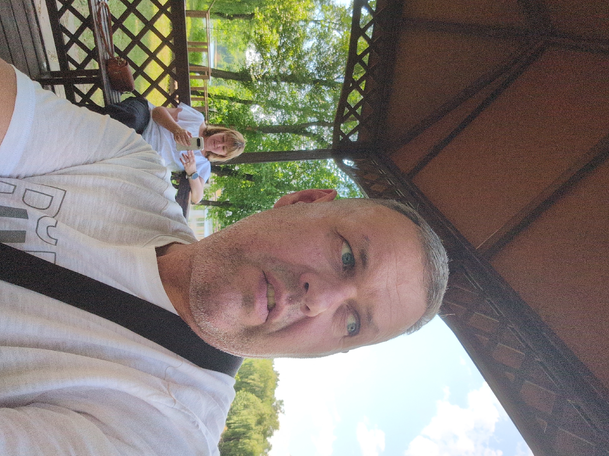 The image shows two people in an outdoor setting, likely a park or recreational area, in Kryvyi Rih, Ukraine. The perspective is a selfie taken by a man in the foreground, who is wearing a white t-shirt with a black strap across it. He has a light beard and is looking towards the camera with a neutral expression.
In the background, a woman is seated on a wooden bench, also wearing a white top. She is holding a smartphone and appears to be engaged with it. A brown handbag is placed on the bench next to her. The setting includes a wooden lattice structure overhead, suggesting a gazebo or covered seating area. Lush green trees are visible behind the bench, indicating a daytime setting, likely summer or late spring given the foliage. The sky is blue with some scattered clouds.
The overall atmosphere appears relaxed and casual, as if the individuals are enjoying a pleasant day outdoors. There is no visible text in the image.