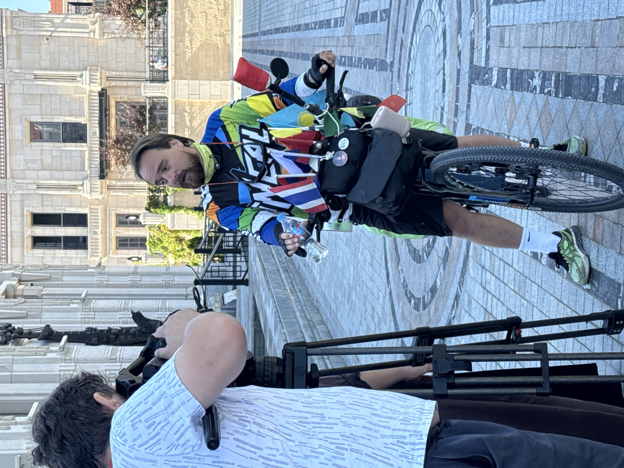 A man with a beard and a colorful cycling jersey stands next to his bicycle, which is adorned with numerous small flags. He is looking to his left, speaking, while a person holding a video camera on a tripod films him. The setting appears to be an outdoor plaza with patterned paving stones and a large, ornate building in the background. The building has classical architectural features and several windows. The man is wearing black shorts, white socks, and athletic shoes. He holds a water bottle in his left hand. The camera person is wearing a white t-shirt with a patterned design. The ground is paved with geometric patterns of light and dark stones, and there are steps leading up to the building. The scene suggests an interview or filming session taking place in a public space.