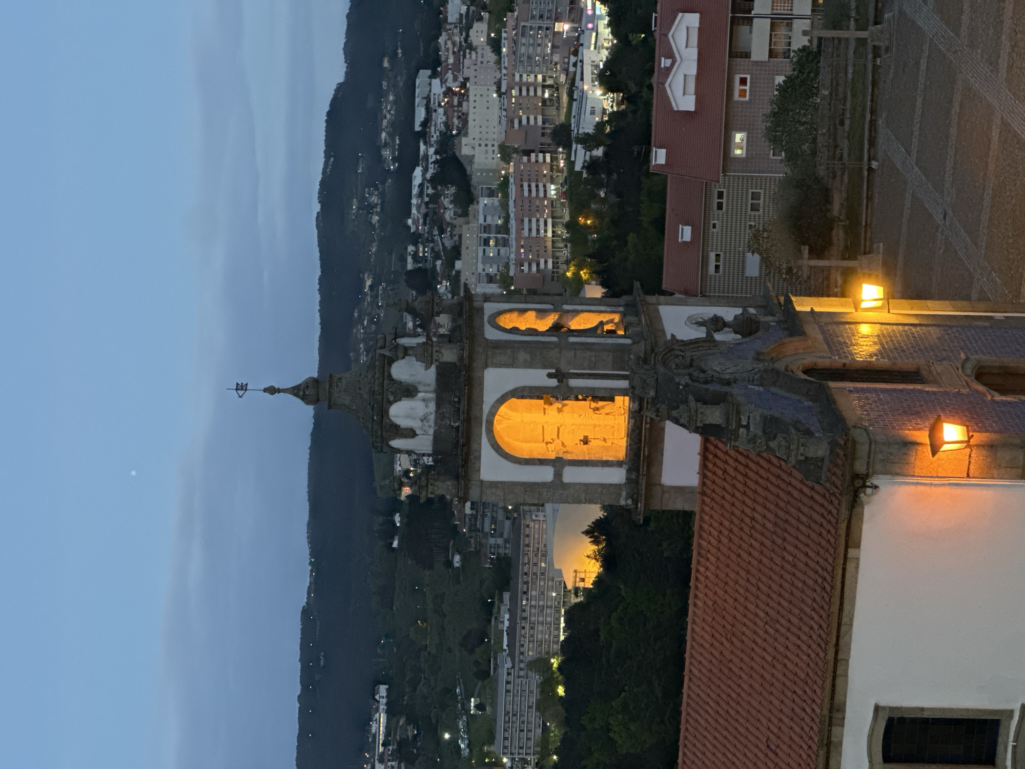 The image is a dusk shot of Vila Real, Portugal, prominently featuring a stone tower illuminated from within. The tower is part of a larger architectural structure, with a red-tiled roof visible. A street lamp is attached to the side of the building, casting a warm glow. In the background, a hillside is dotted with buildings and trees, all visible under a twilight sky. The overall atmosphere is tranquil and scenic.