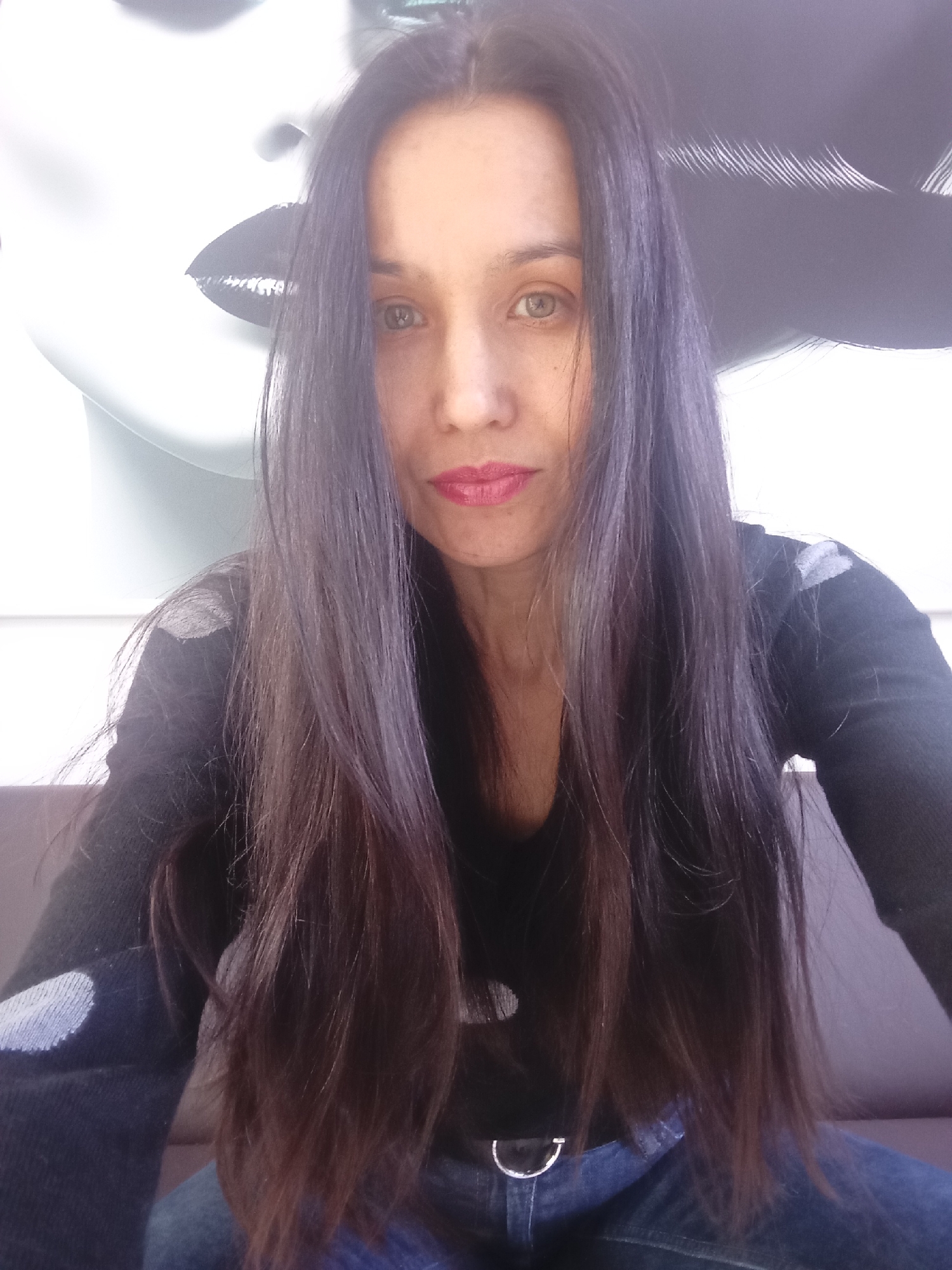 A woman is shown in a selfie-style, close-up portrait. She has long, dark hair, red lipstick, and is wearing a dark-colored shirt or sweater with light-colored polka dots on the sleeves. She appears to be wearing jeans. In the background, partly visible, is what seems to be a white sculptural or art piece depicting a face, with prominent lips. There is no explicit visual evidence linking this image to Izhevsk, Russia. There are no discernible actions or interactions taking place in the image, only the woman's direct gaze towards the camera.