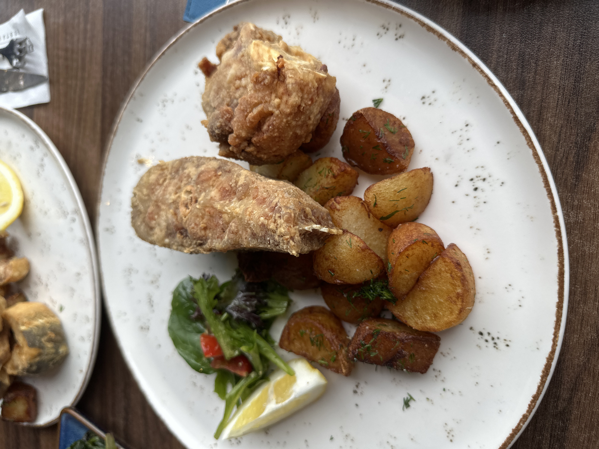The image shows a plate of food on a wooden table. The plate contains two pieces of fried food, possibly chicken, a portion of roasted potatoes seasoned with herbs, a small salad with a slice of tomato, and a wedge of lemon. A portion of another plate of food is visible on the left side of the image. The scene appears to be in a restaurant setting in Vidin, Bulgaria.