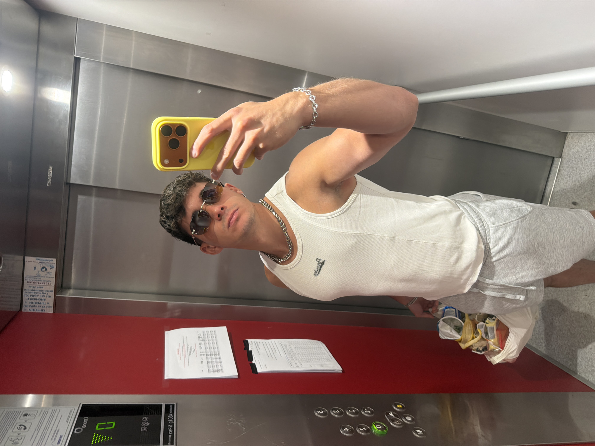 A young man is taking a selfie in an elevator. He is wearing a white tank top, gray shorts, sunglasses, and a silver bracelet. He is holding a yellow phone. In the background, the elevator walls are stainless steel and red. There are papers and a bag of trash on the floor. The elevator buttons are visible. The time of day is not discernible. The location is Sofia, Bulgaria.