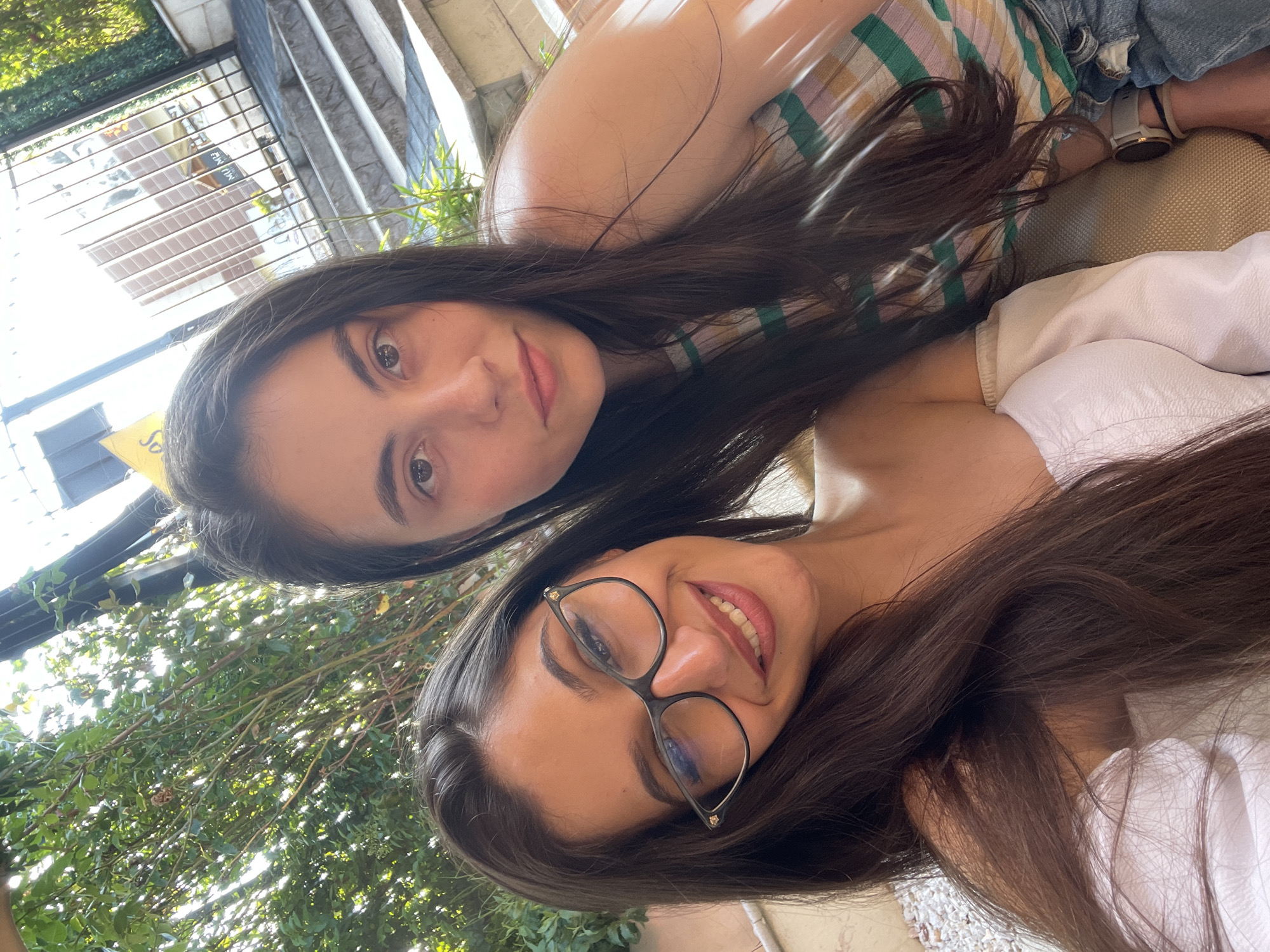 The image captures two young women in a selfie-style close-up, set outdoors in Pazardzhik, Bulgaria, under bright daylight. The woman positioned closer to the camera on the left has long dark brown hair and a smiling expression. She wears black-framed eyeglasses with gold-colored embellishments and a white off-the-shoulder top. The woman on the right, positioned slightly behind and to the side, also has long dark brown hair and maintains a neutral expression while looking directly at the camera. She is dressed in a horizontally striped top featuring green, pink, yellow, and white bands, along with blue denim jeans. A light-colored smartwatch with a grey or white band is visible on her left wrist.
The background behind the subjects consists of dense green foliage on the left, with bright sunlight filtering through the leaves. To the right, a dark metal fence or gate is prominent, beyond which a multi-story building with reflective window surfaces and a partially visible yellow sign with dark text can be discerned. Below the fence, a series of light-colored concrete steps ascends diagonally. White pebbles or gravel are visible on the ground in the lower-left section of the frame. The women appear to be seated or leaning against a light brown or tan textured surface.