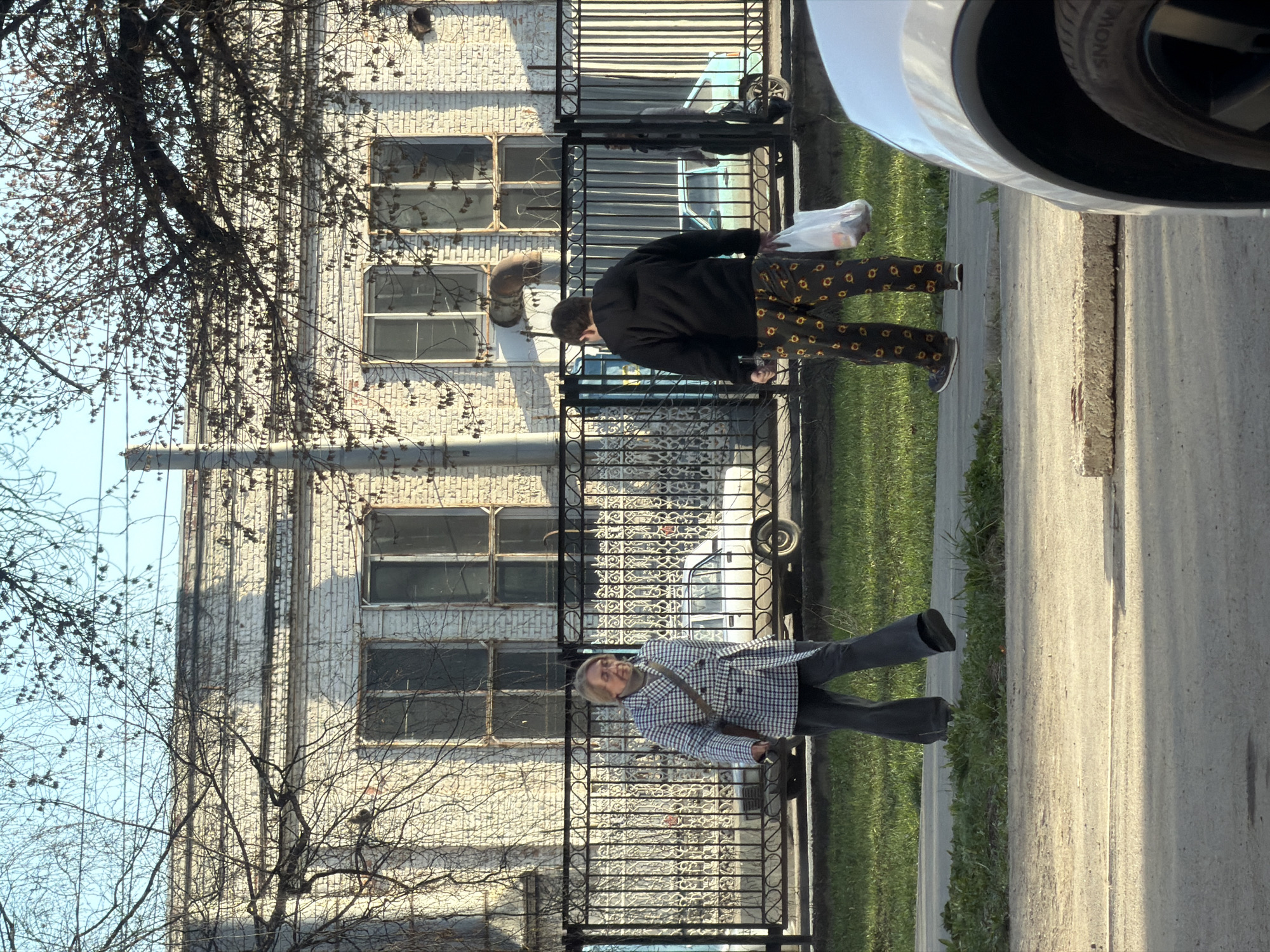 This media file captures an outdoor street scene in Izhevsk, Russia, under bright daylight. The image is oriented sideways, but depicts a sidewalk running horizontally at the bottom, adjacent to a decorative black metal fence. Beyond the fence stands a light-colored, possibly white or pale grey, brick building with multiple rectangular windows, some of which appear worn. Bare tree branches, characteristic of late autumn, winter, or early spring, are prominent in the foreground on the left, with a clear blue sky visible behind them.
Two individuals are walking along the sidewalk. The person closer to the foreground, on the left, appears to be an older individual with light-colored hair, wearing a distinctive blue, white, and black checkered jacket, dark trousers, and carrying a shoulder bag. They are walking towards the right side of the frame, with their face partially visible. The second person, positioned further back and to the right, has dark hair and is dressed in a black top or jacket paired with unique dark trousers adorned with a repeating yellow or orange circular pattern. This individual is holding a white plastic bag in their left hand and is also walking towards the right, with their back mostly turned to the viewer.
Visible objects include a partial view of a white car's front wheel and fender in the upper right corner, with the word "SNOW" partially legible on the tire, suggesting it might be a snow tire or all-season tire. Behind the black fence, a light blue car is also partially visible. Metal pipes or conduits run vertically on the building's facade. The sidewalk is made of concrete, and a strip of green grass separates it from what appears to be a curb or another section of the road. The sunlight casts distinct shadows, indicating a clear, sunny day. No strong emotions are discernible from the individuals.