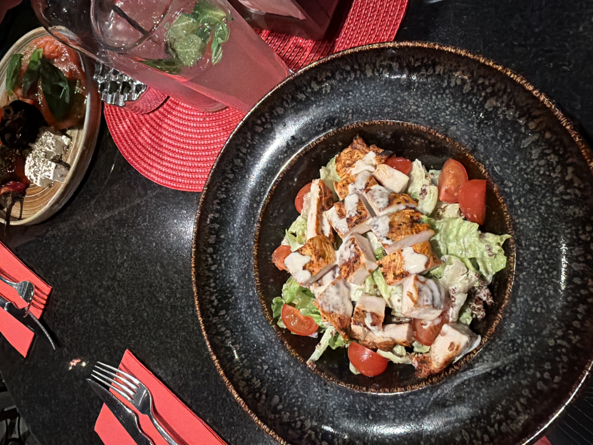 A high-angle shot presents a table setting with a black speckled bowl containing a salad, accompanied by a drink, appetizer, and cutlery. The salad, situated in the center of the frame, features lettuce, tomato halves, and sliced grilled chicken drizzled with a white sauce. A red drink with ice and mint leaves sits on a red placemat in the upper left of the frame, adjacent to the bowl. To the left of the drink is a plate with an appetizer composed of tomatoes, greens, dark-colored food (possibly olives or chocolate), and white cheese. In the lower left corner, two forks and a knife, each resting on a red napkin, complete the setting. The table is a dark, possibly black, material. The specific location in Sofia, Bulgaria, cannot be ascertained from the image alone.