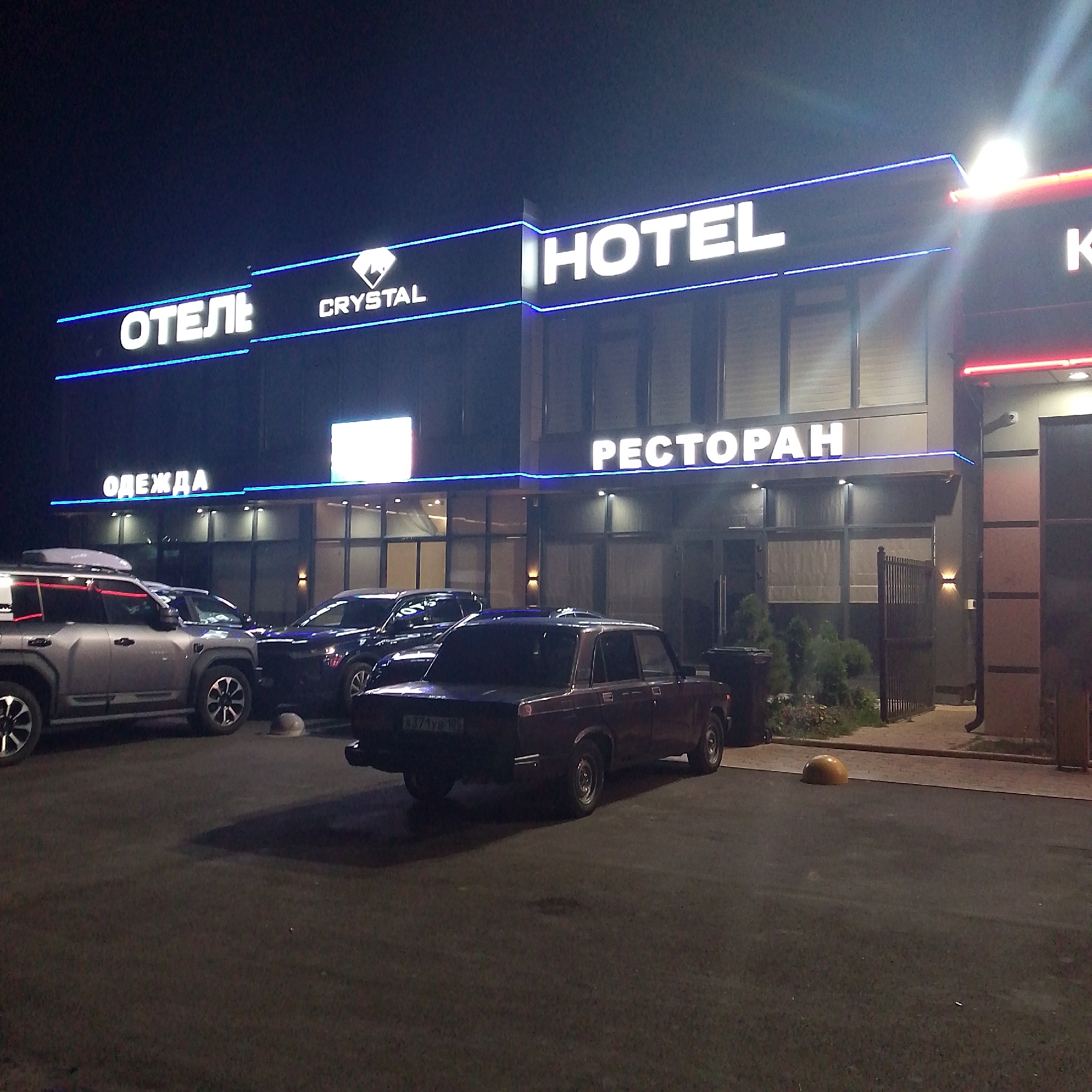 The image shows a hotel and restaurant complex at night. The building features prominent illuminated signage. On the left, the Russian word "ОТЕЛЬ" (Hotel) is displayed in white illuminated letters, followed by a logo and then "CRYSTAL". To the right, the English word "HOTEL" is displayed in large white illuminated letters. Further to the right, the Russian word "РЕСТОРАН" (Restaurant) is also in illuminated white letters. A red illuminated sign is visible on the far right.
In the foreground, several cars are parked. A silver SUV with roof rails is on the left. Next to it is a dark-colored sedan, and to the right of that is a burgundy-colored sedan with a visible license plate reading "A371УЕ 102". The ground appears to be asphalt, and there are some small plants and a fence visible behind the cars. The overall environment suggests an outdoor area adjacent to the building, likely a parking lot. The lighting is artificial, coming from the building's signs and some wall-mounted lights. There are no people visible in the image. The location is specified as Krasnooktyabrskoye, Russia.