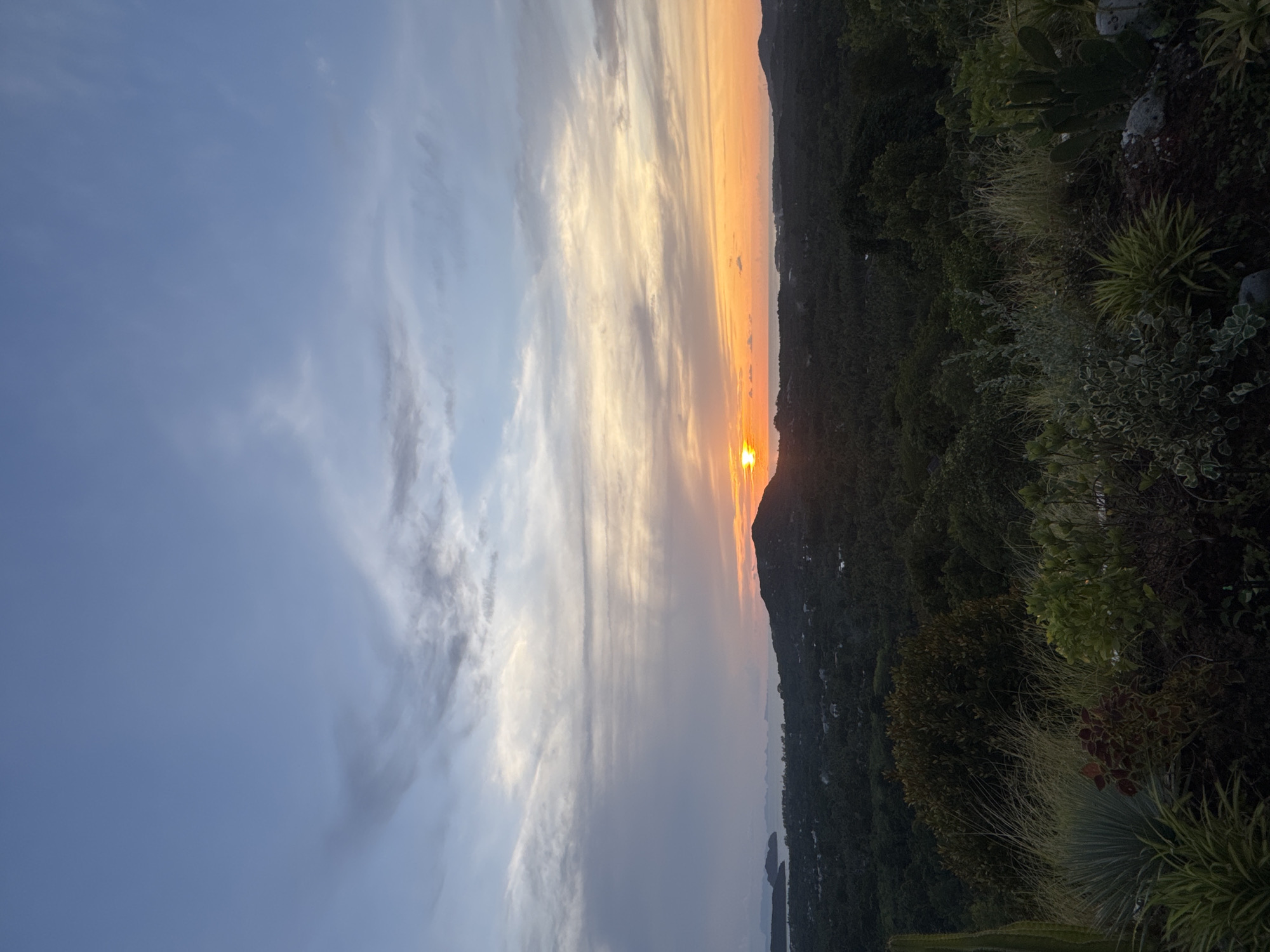 The image presents a view of a sunset over Baan Nai Suan, Thailand. The sky transitions from light blue with wispy clouds on the left to vibrant orange and yellow hues near the horizon on the right. The sun, a bright, circular orb, is partially obscured by a dark, silhouetted mountain range. A lush, green landscape, likely consisting of vegetation, foliage, and trees, fills the foreground.