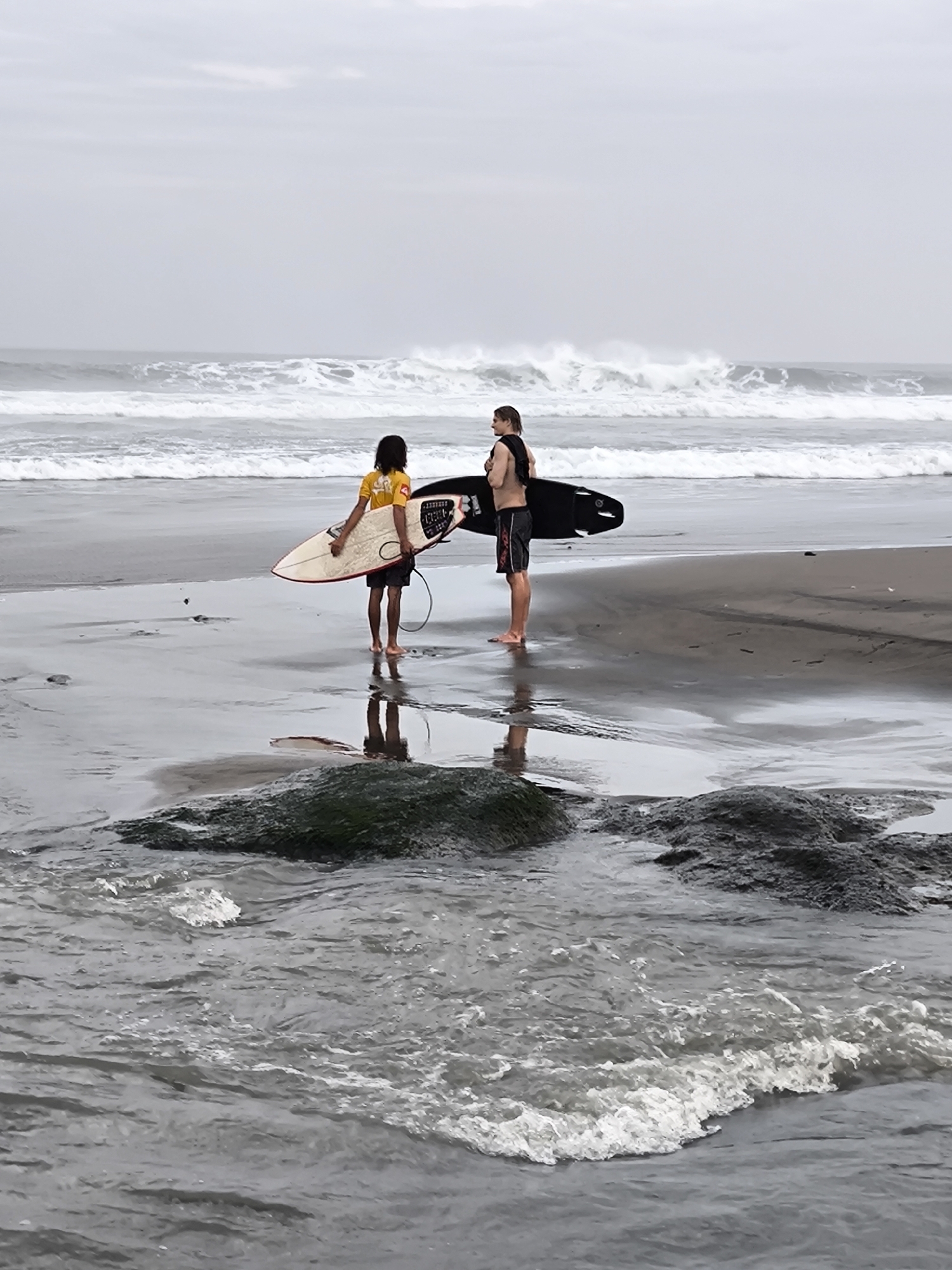 Two surfers stand on a wet sandy beach in front of the ocean, holding their surfboards. The surfer on the left is a child wearing a yellow t-shirt and dark shorts, holding a light-colored surfboard. The surfer on the right is an adult wearing dark shorts and a black shirt draped over their shoulders, holding a black surfboard. The ocean behind them has large waves with white caps. The sky is overcast. The location is the beach in Mengwi, Indonesia.