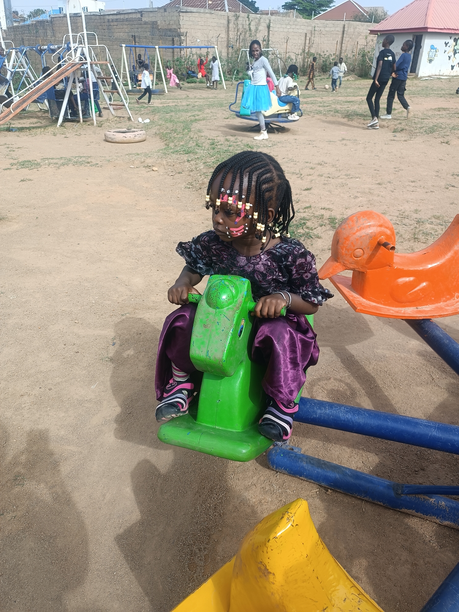 A medium shot captures a young girl with dark hair adorned with beads and face paint sitting on a green, horse-shaped, spring-mounted playground ride. She is wearing a purple dress and black shoes with pink and white stripes. The ride is part of a roundabout carousel, visible with faded yellow and orange plastic animals.
In the background, other children are playing in a playground setting with swings, a slide, and a small merry-go-round. The background includes a building with a red roof and a painted facade, as well as a brick wall topped with barbed wire. The ground appears to be a dirt or sandy surface. The scene is located in Jos, Nigeria.